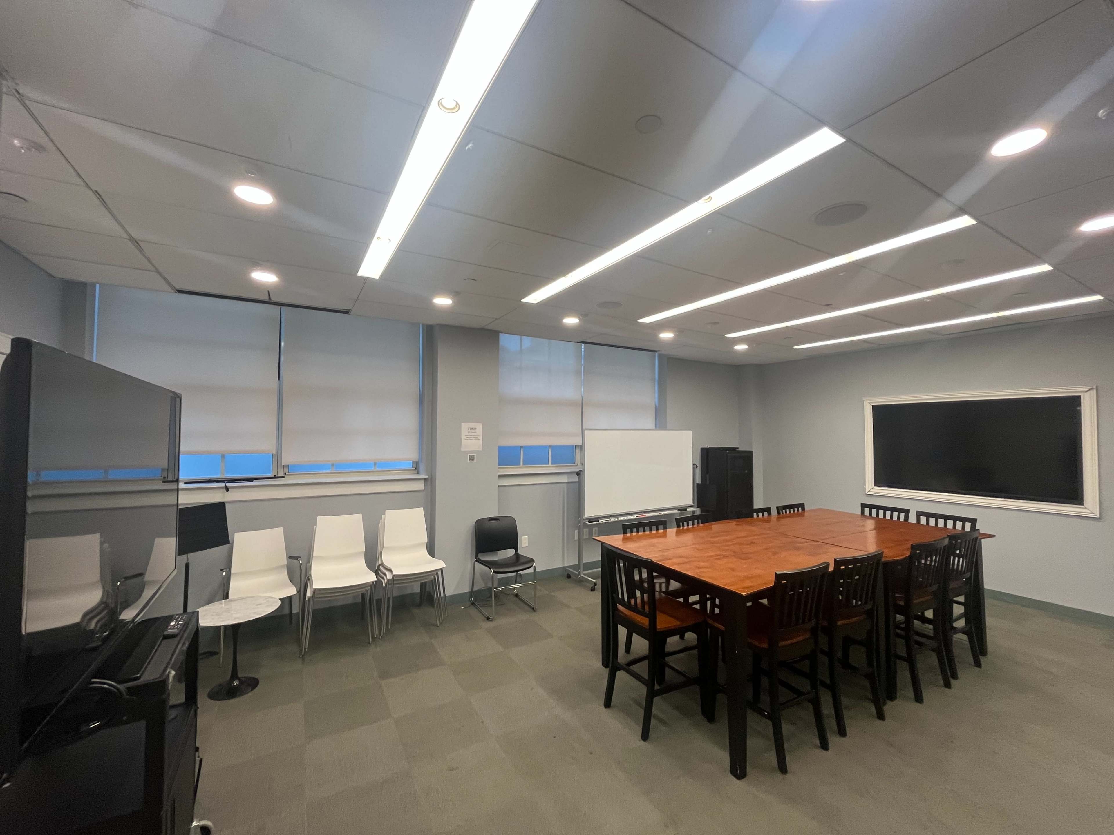 The image shows a modern conference room with a large wooden table, several chairs, and two large screens on the walls.
