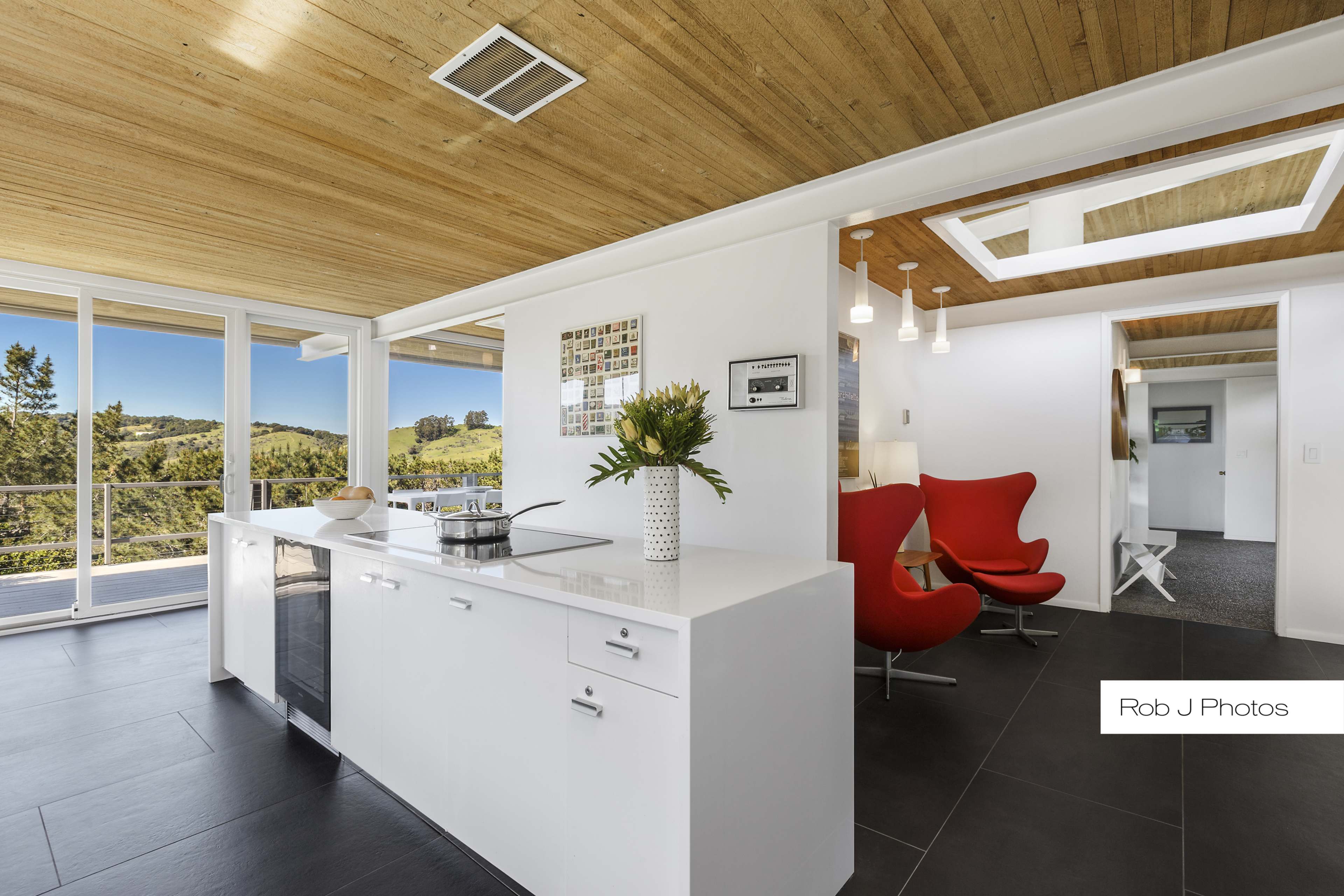 A modern kitchen with a white island, dark tile flooring, and large windows offering a view of greenery.