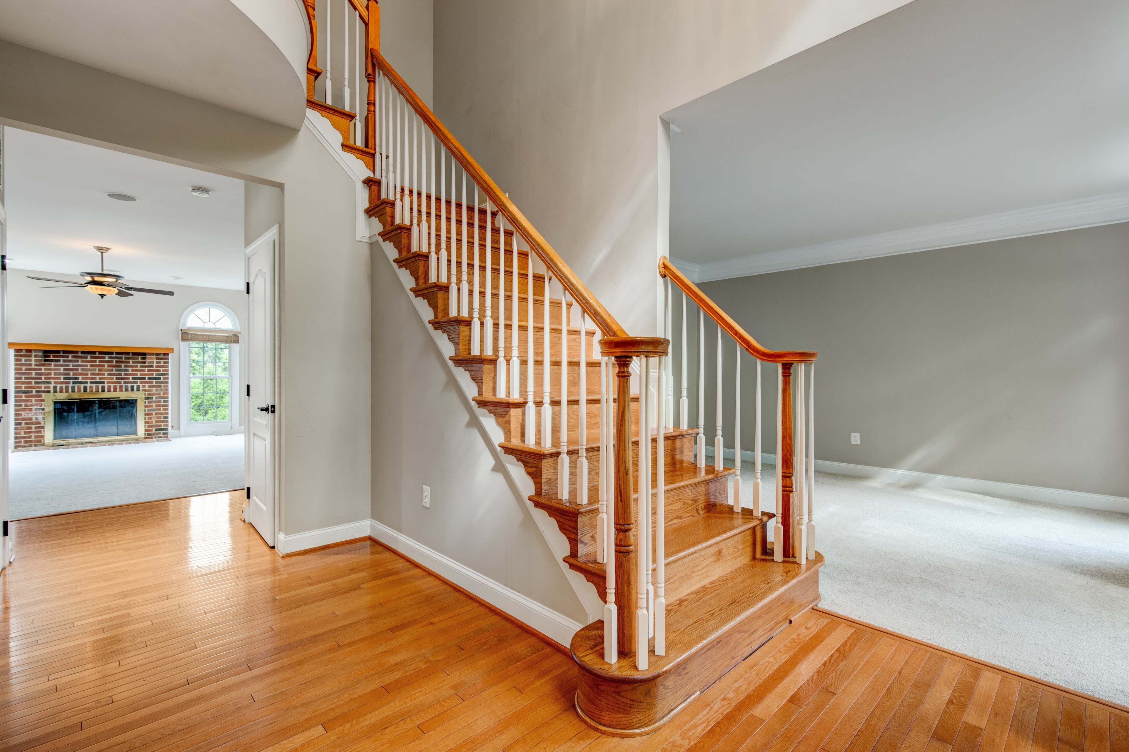 A wooden staircase leading to an upper level in a well-lit hallway with hardwood flooring and an archway leading to another room.
