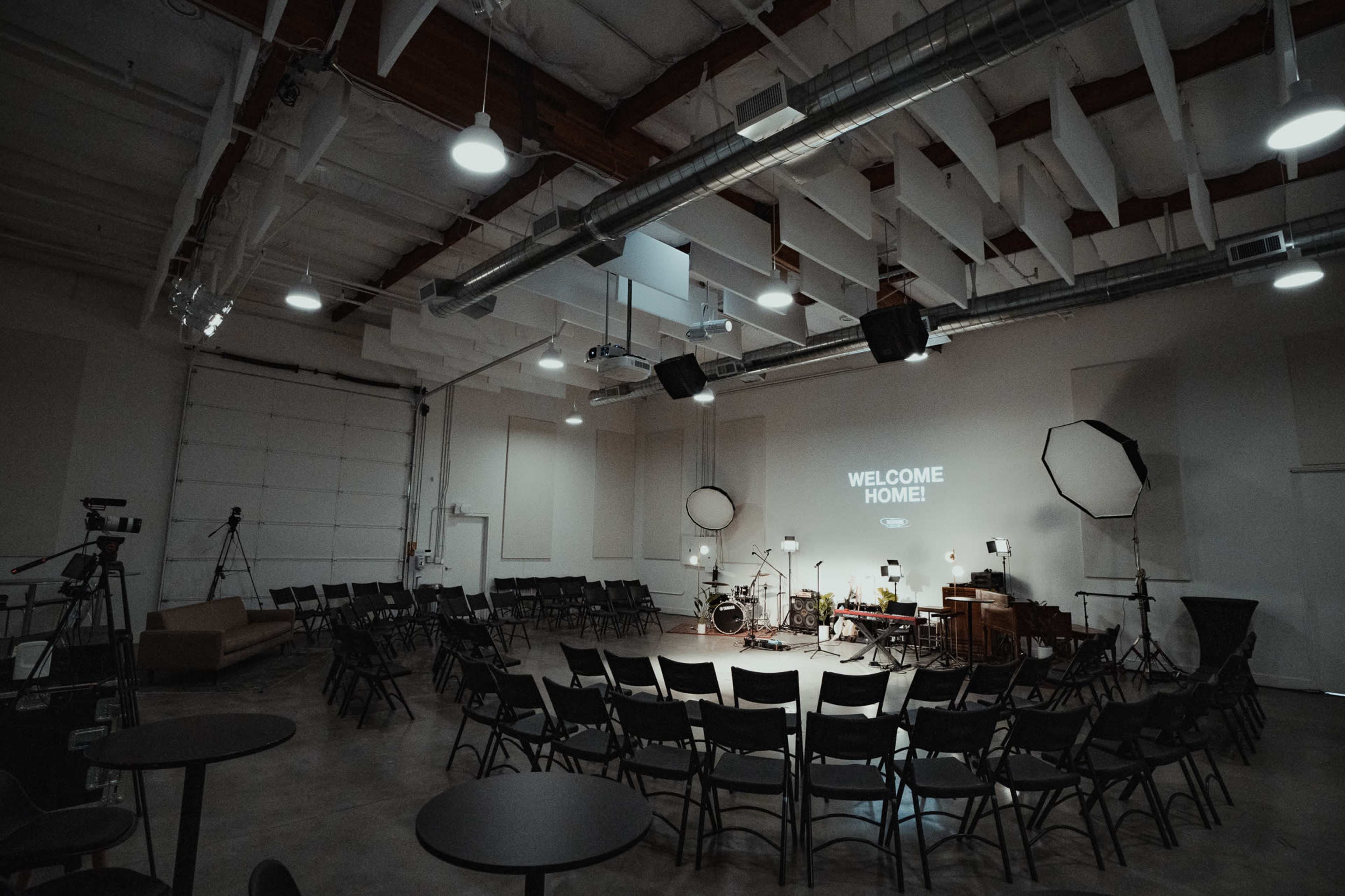 A spacious room is set up with circular rows of chairs facing a stage featuring a projector screen that reads "WELCOME HOME!" alongside musical instruments and lighting equipment.