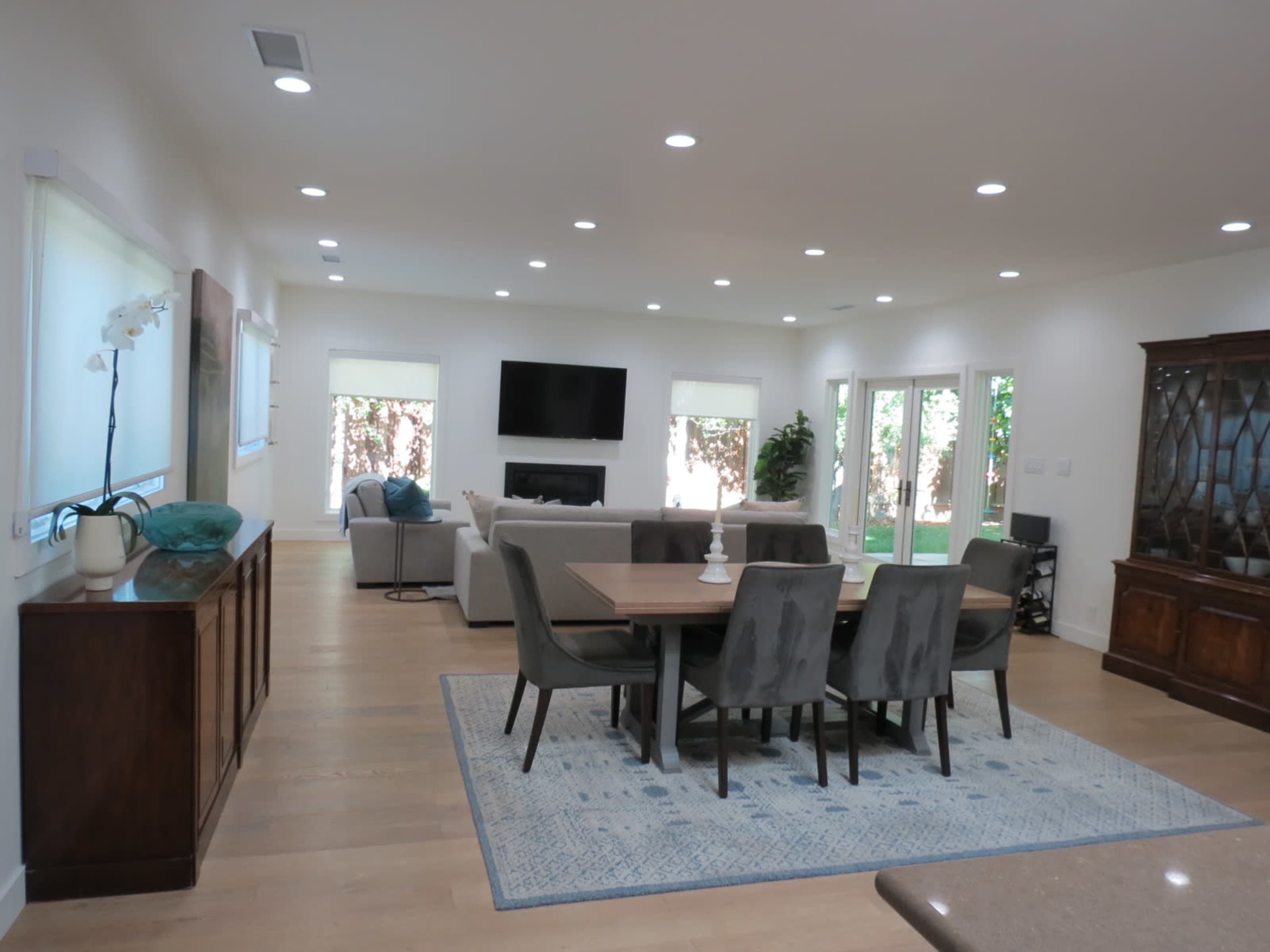 The image shows a modern, open-concept living and dining area featuring a dining table surrounded by chairs, a lounge area with a couch, and large windows leading to a backyard.