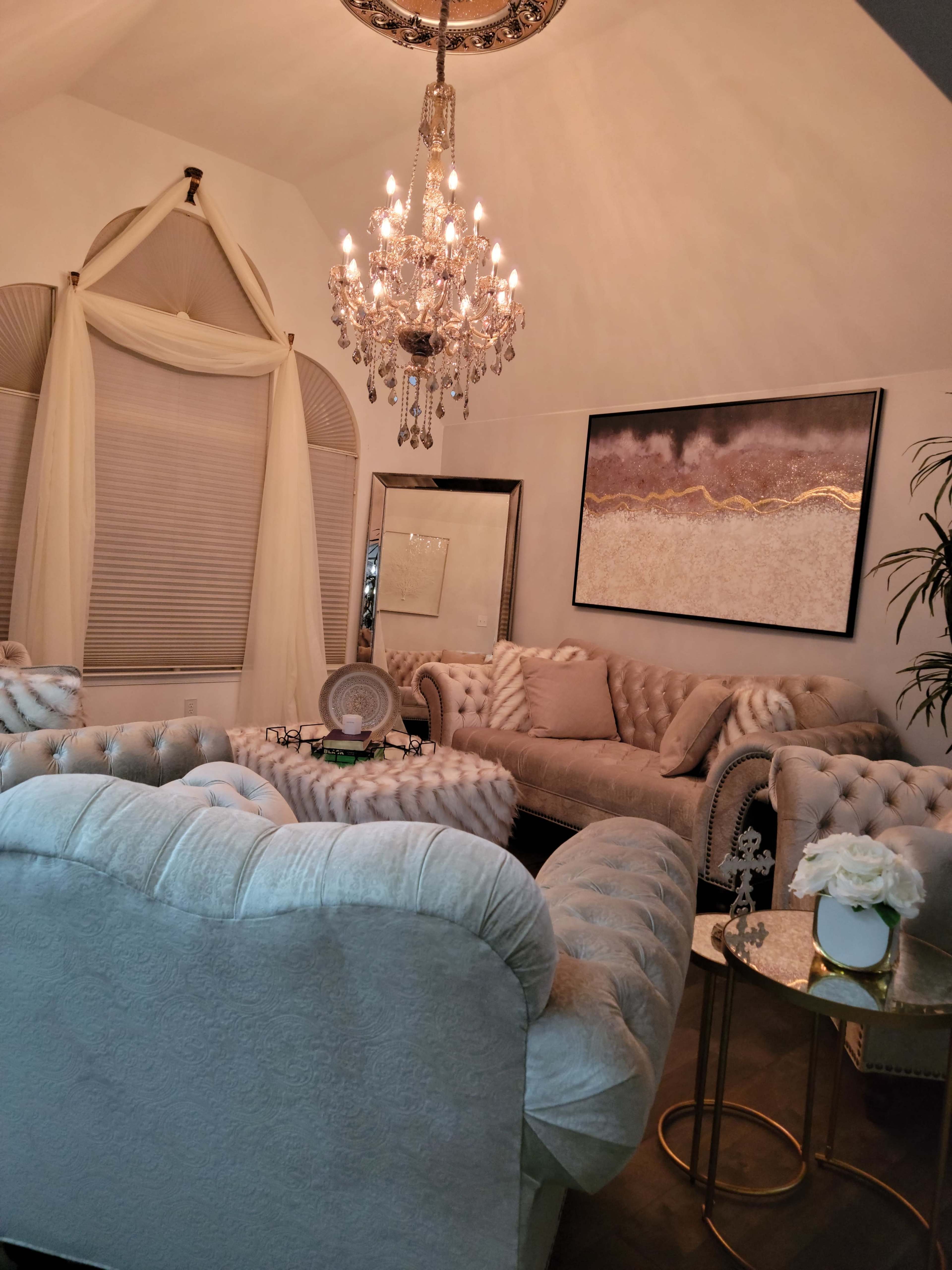 A elegantly furnished living room featuring plush sofas, a chandelier, and decorative elements including a mirror and a small table with floral arrangements.