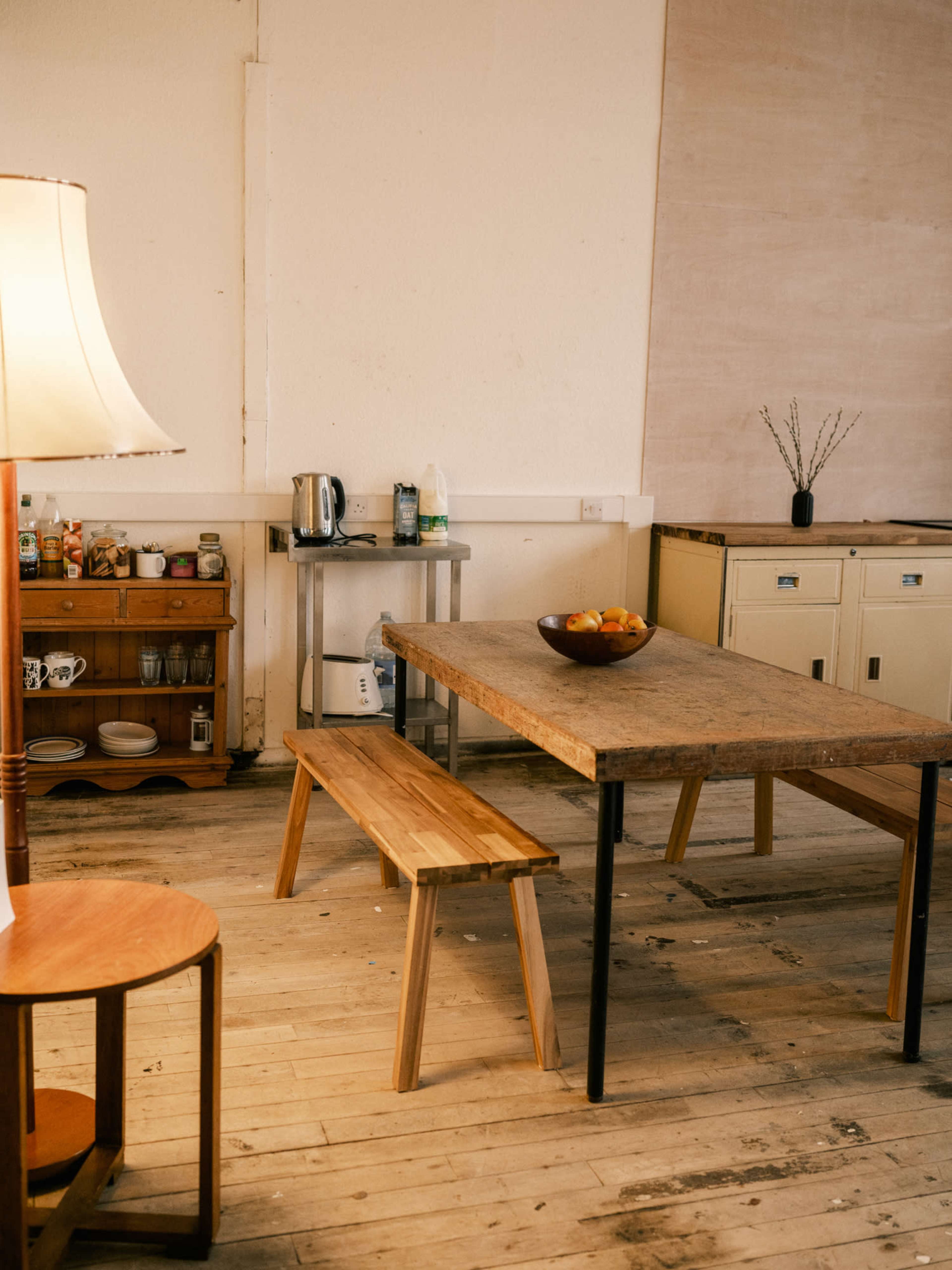 The image shows a simple kitchen space with a wooden table and bench, a kettle on a stand, a bowl of fruit on the table, and shelves with various kitchen items in the background.