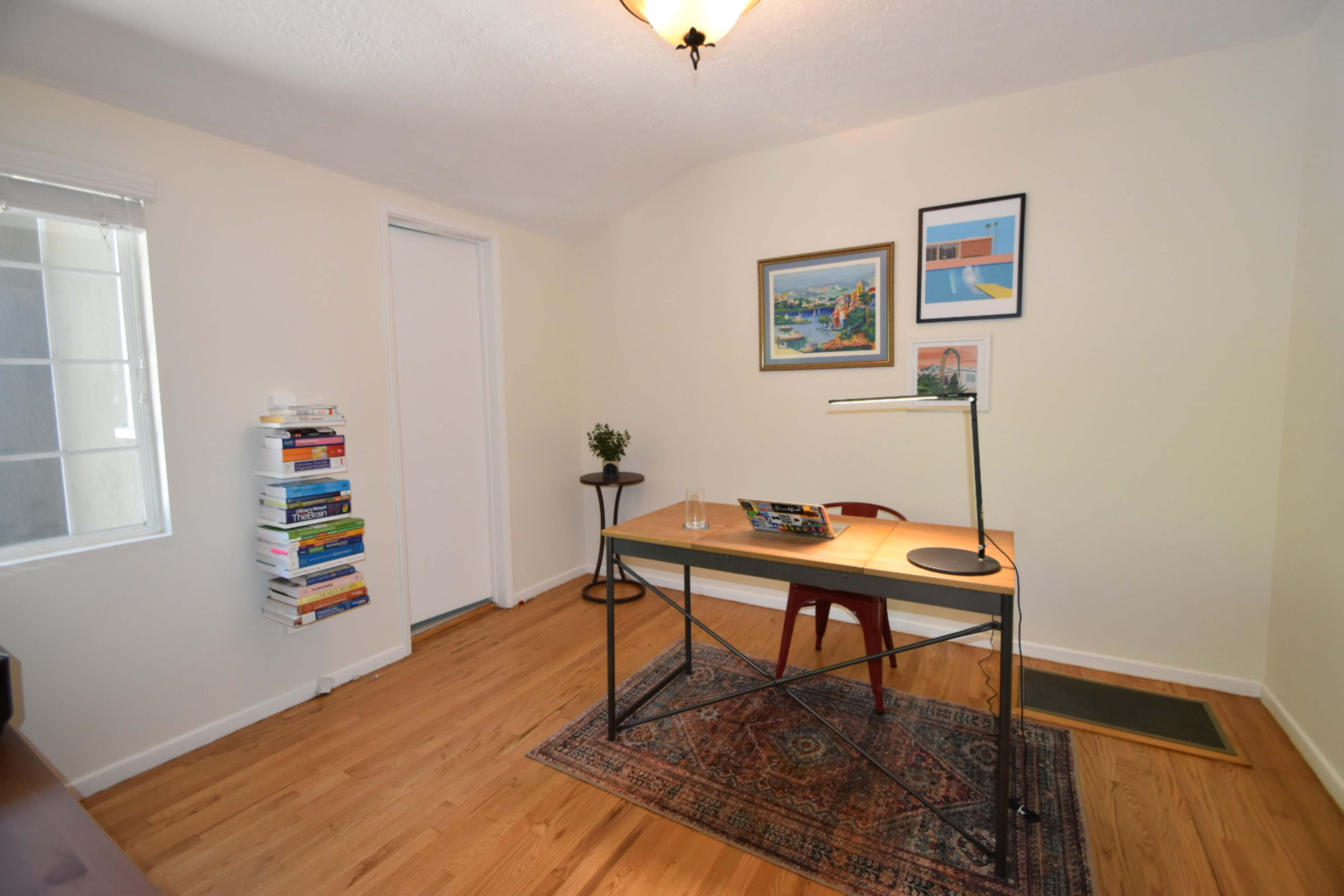 The image shows a minimalist home office with a wooden desk, a chair, a lamp, and a small bookshelf stacked with books.
