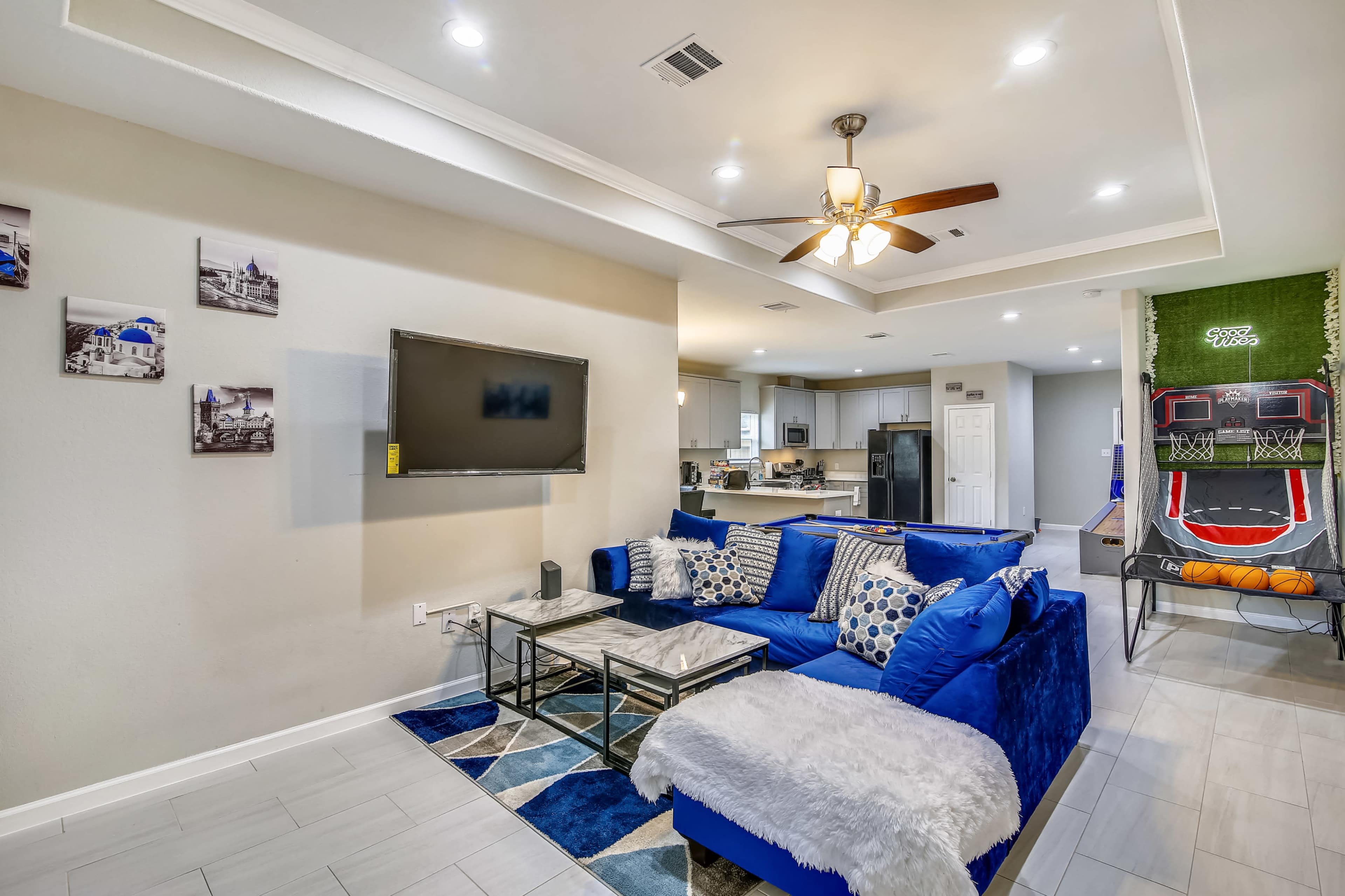 A modern living room featuring a blue sectional sofa, a flat-screen TV mounted on the wall, and a kitchen visible in the background, along with a basketball shooting game in the corner.