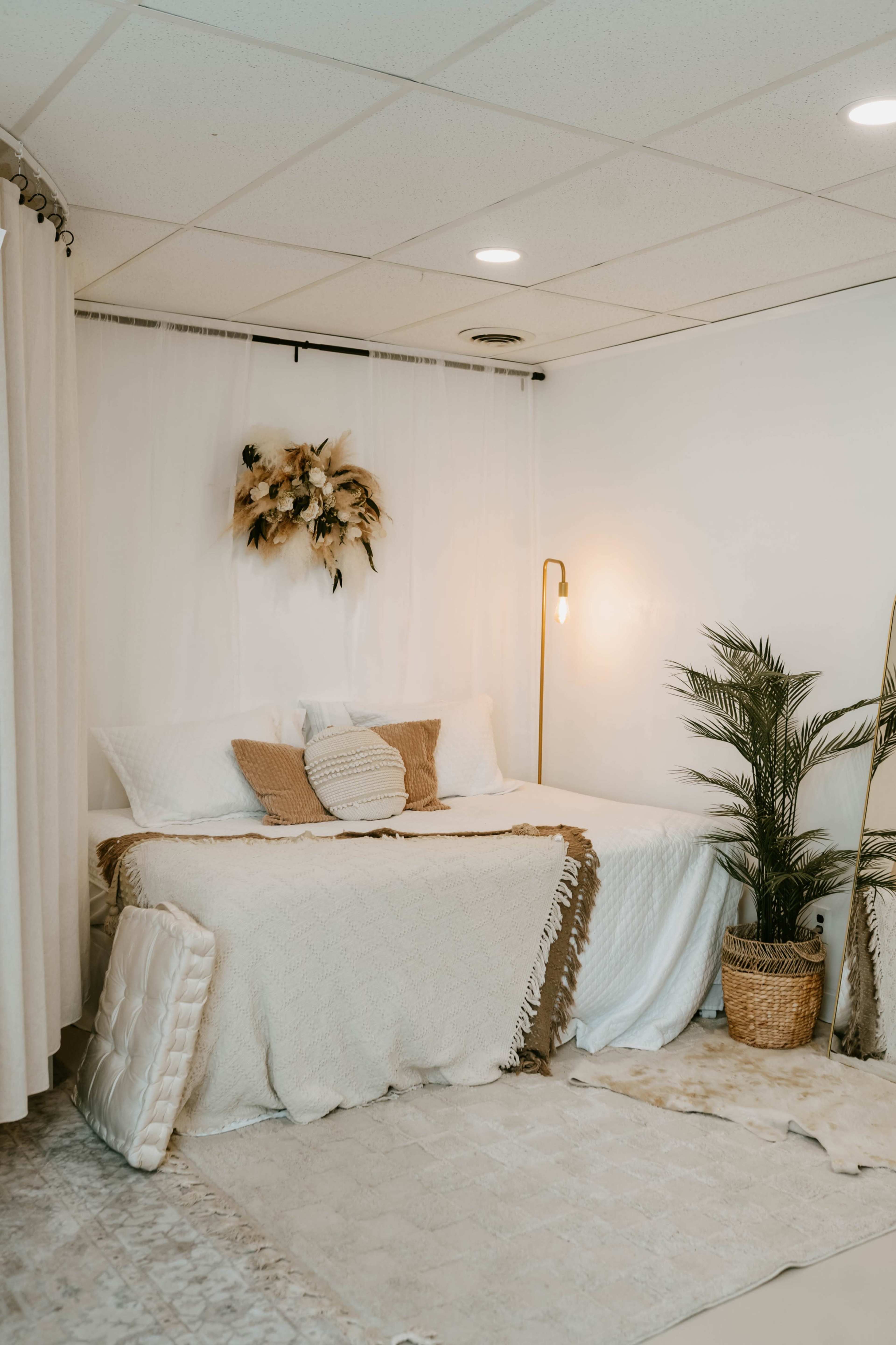 The image shows a neatly arranged bedroom with a bed covered in neutral bedding, a decorative plant, and a floor lamp, all set against a light-colored background.