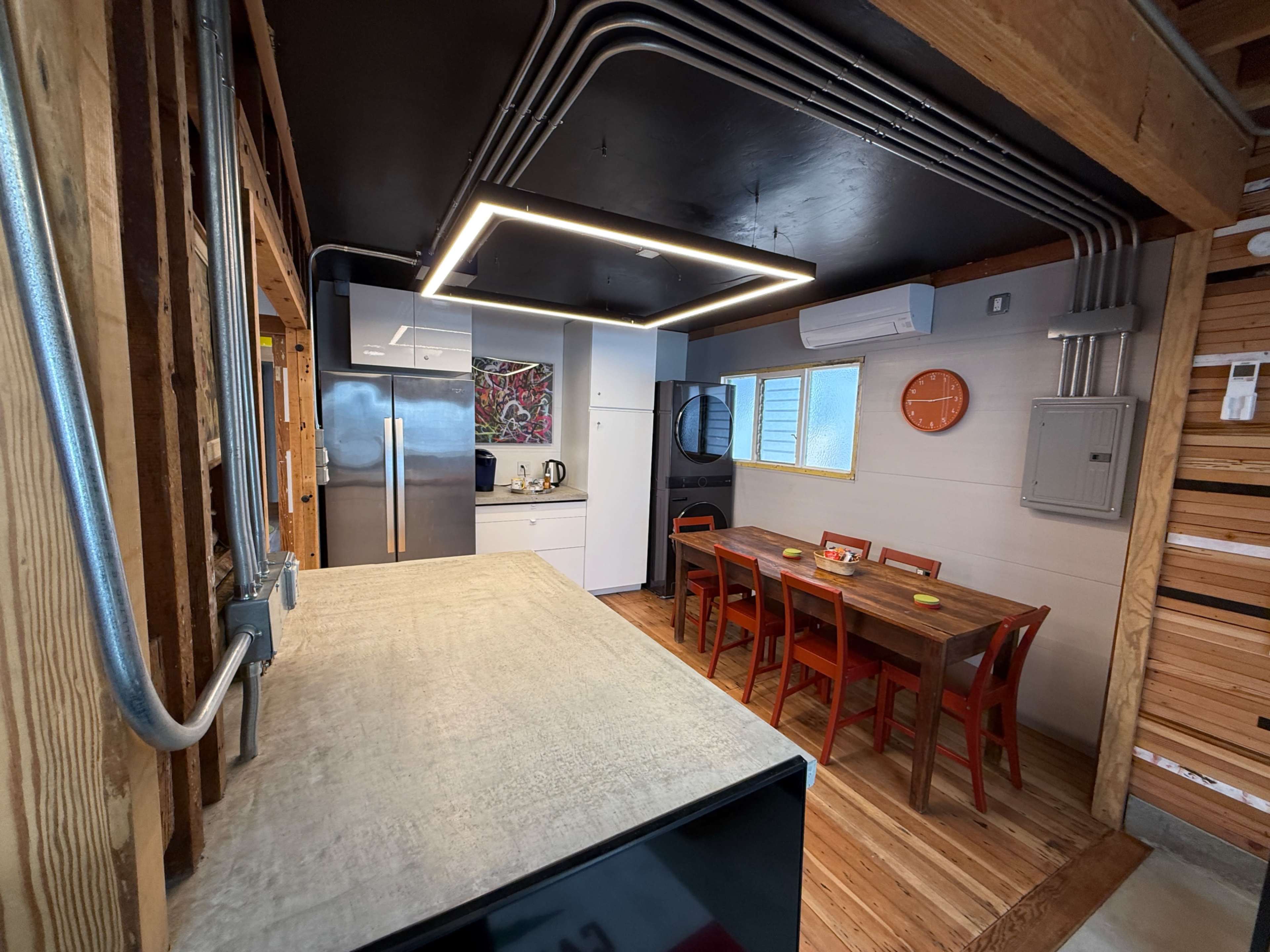 The image shows a modern kitchen and dining area with stainless steel appliances, a wooden dining table, and exposed piping along the ceiling.