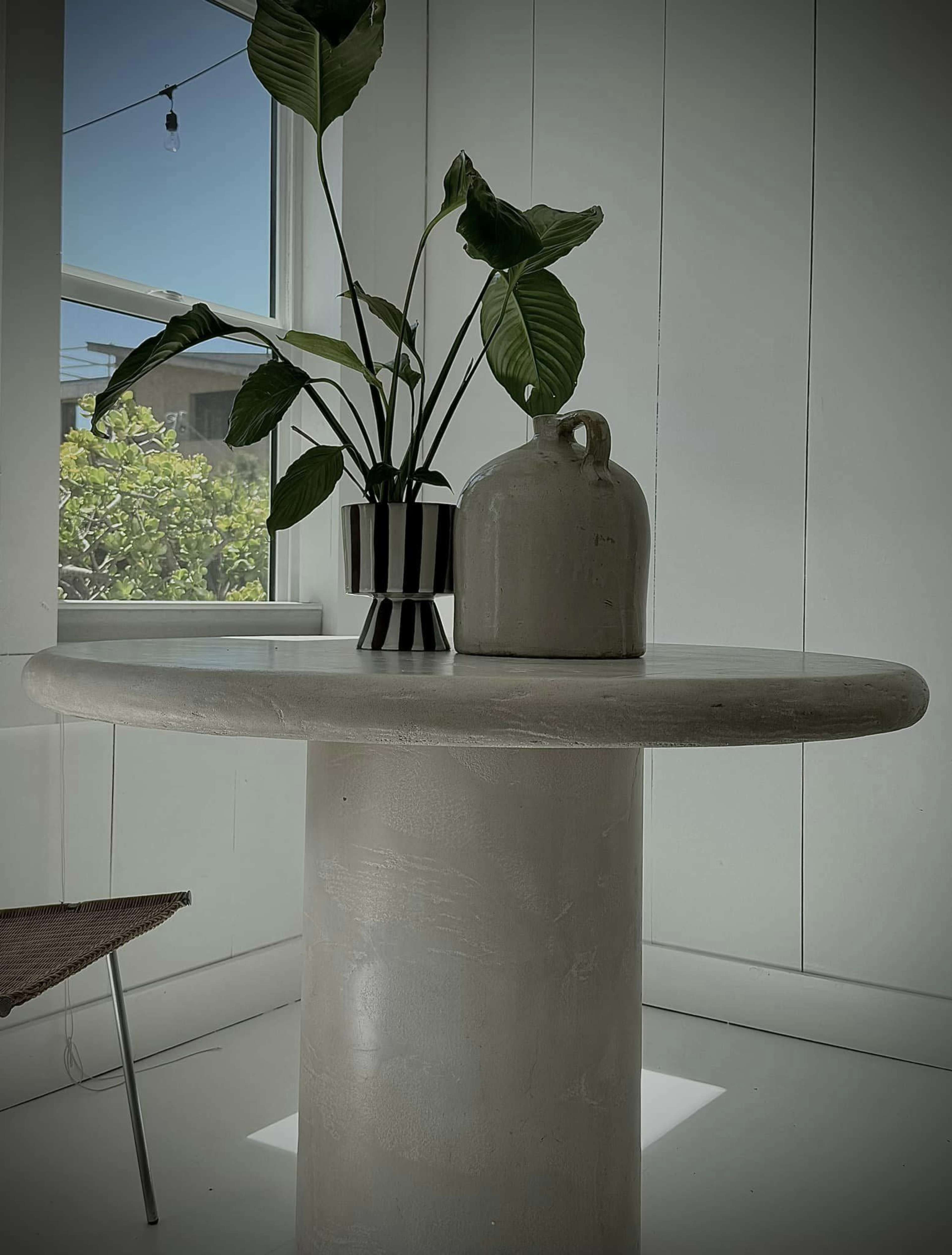 A round table with a smooth surface holds a plant in a vase and a ceramic jug, set against a bright, minimalistic interior with large windows.
