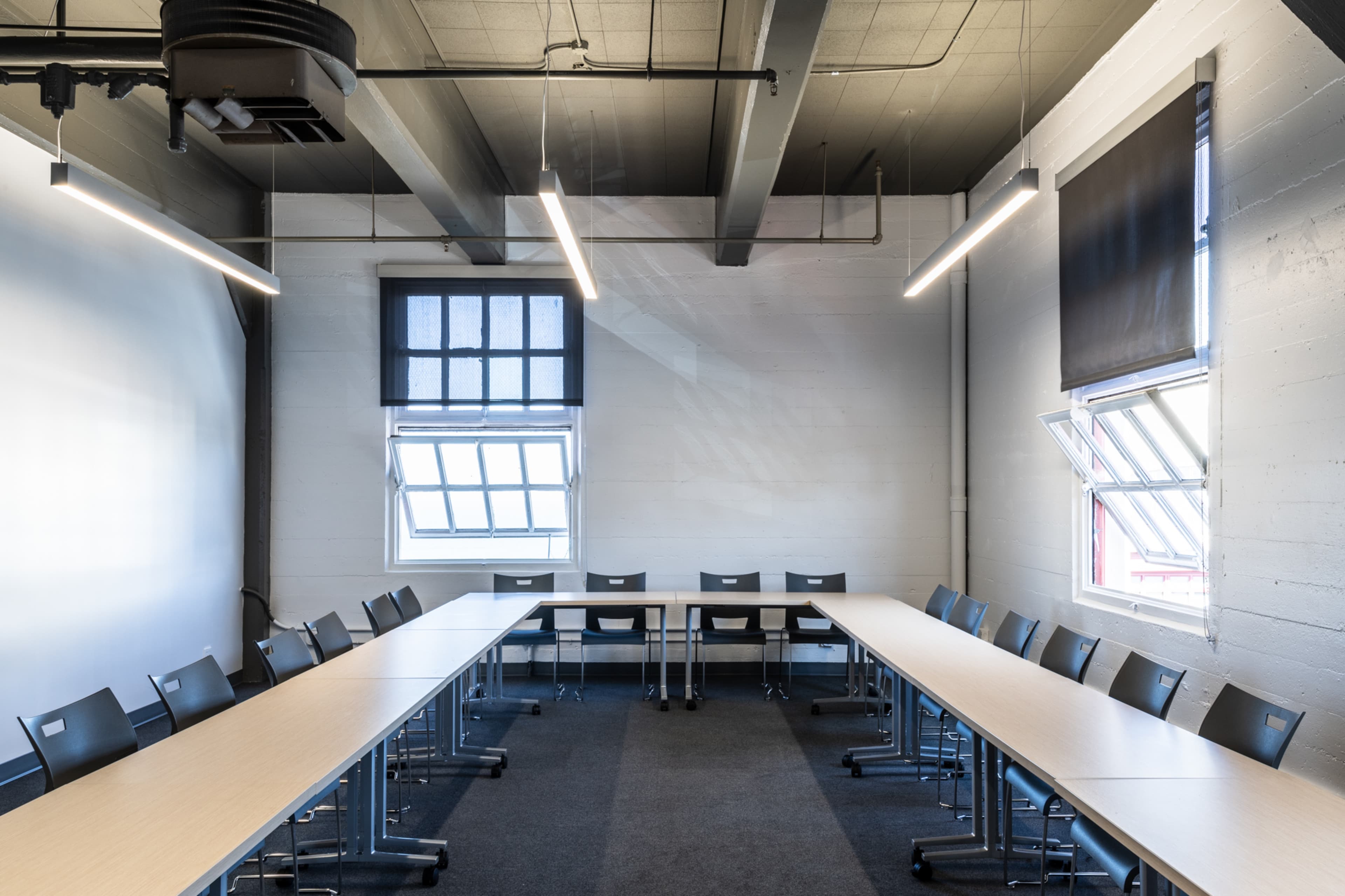 A spacious meeting room features two long tables arranged in a U-shape, surrounded by black chairs, with windows allowing natural light and minimalistic lighting overhead.