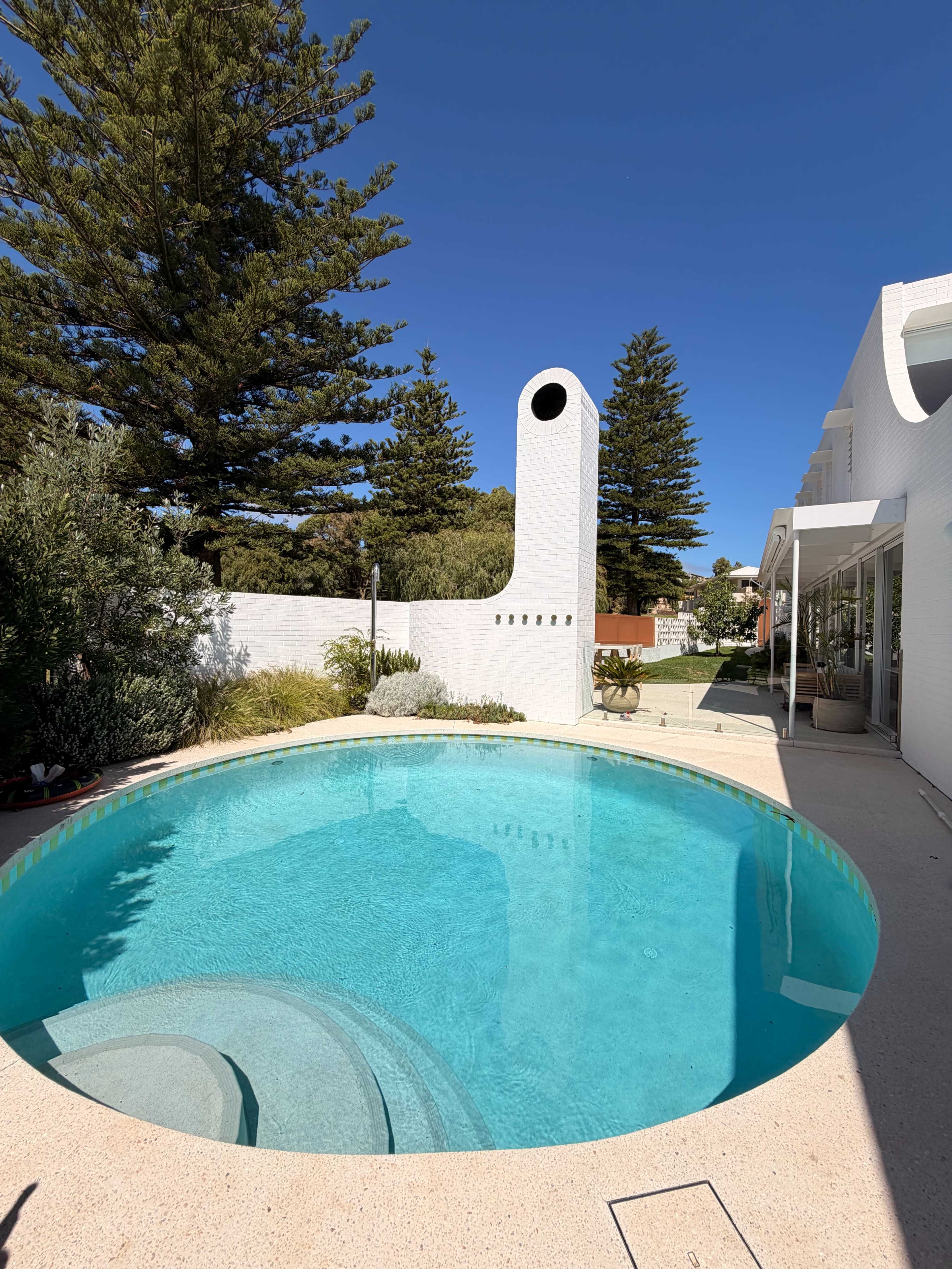 The image shows a circular swimming pool surrounded by a well-maintained garden, with a tall white chimney and clear blue sky in the background.