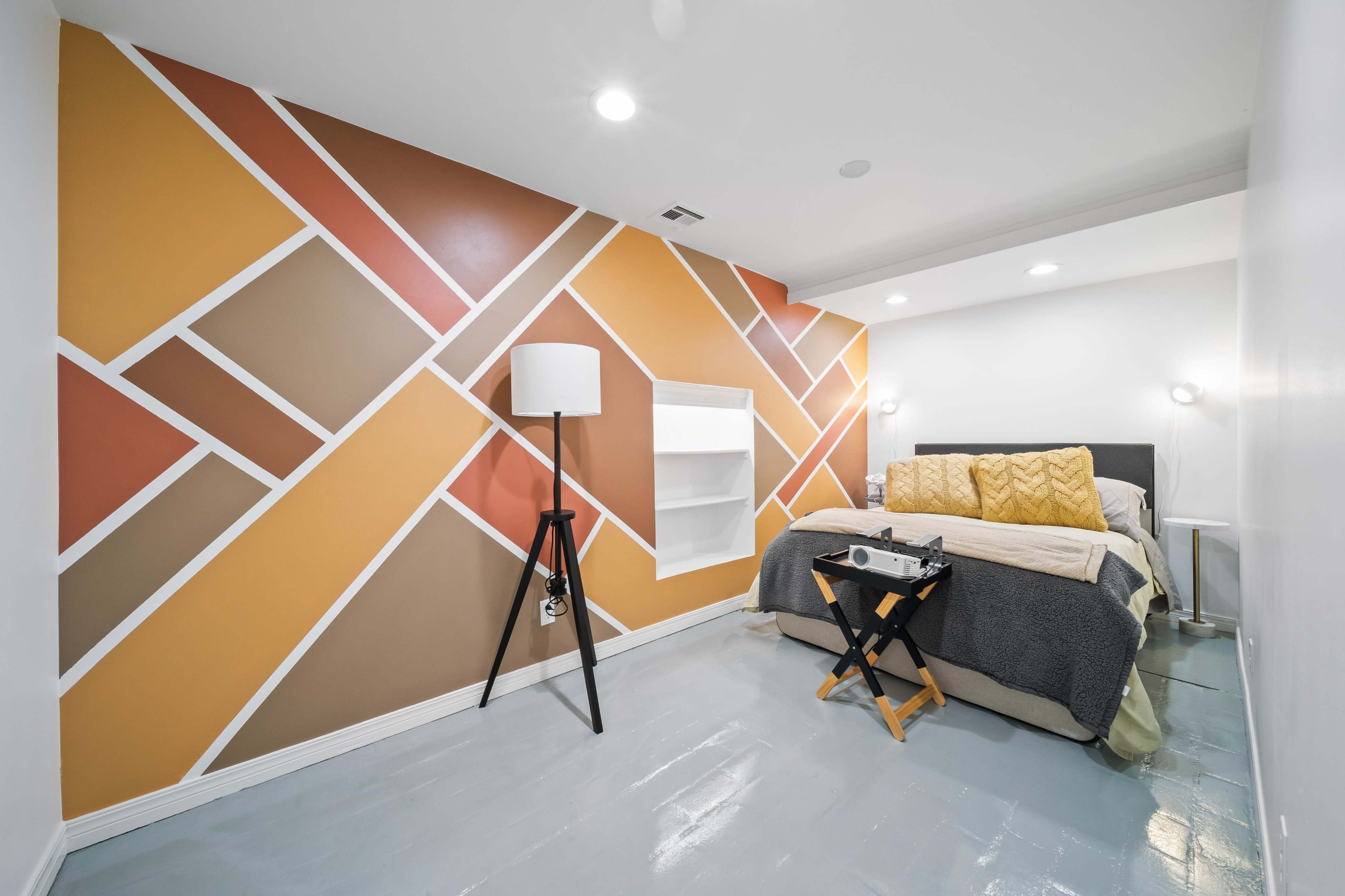 The image shows a modern bedroom with a colorful geometric accent wall, a bed with decorative pillows, and a bedside lamp on a tripod stand.