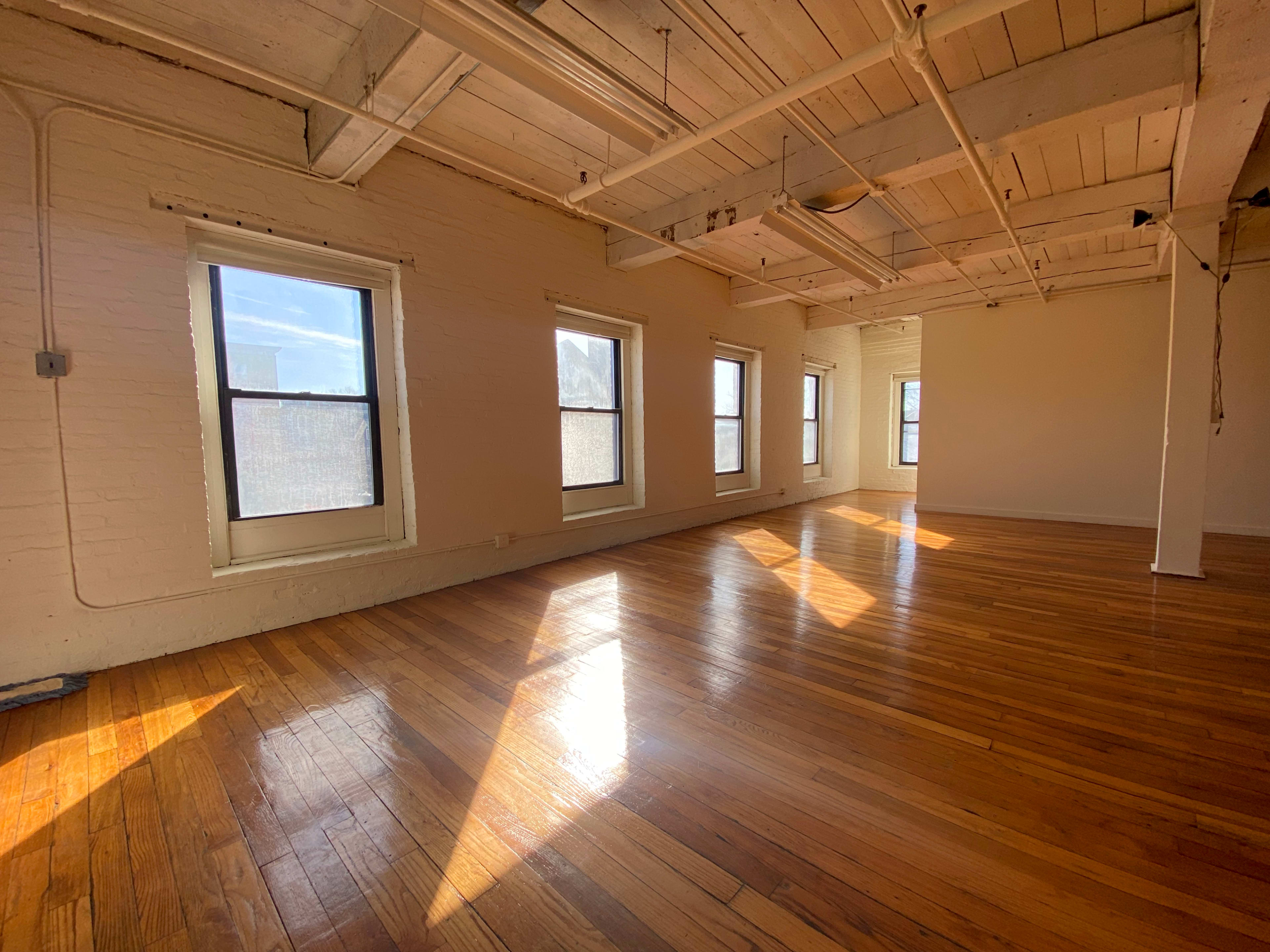 A spacious, empty room with hardwood floors and multiple windows allowing natural light to enter.