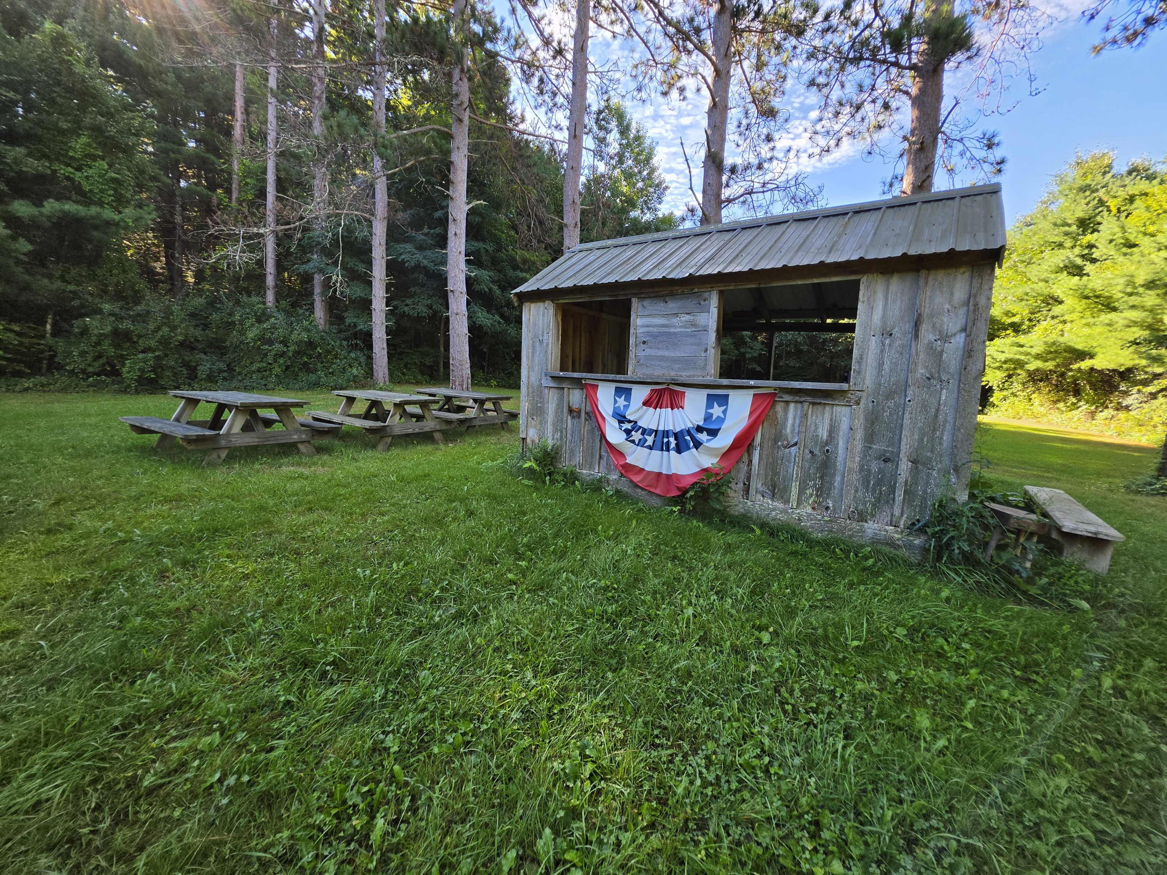 The image shows a wooden kiosk with a decorative red, white, and blue banner in a green, grassy area surrounded by trees and picnic tables.