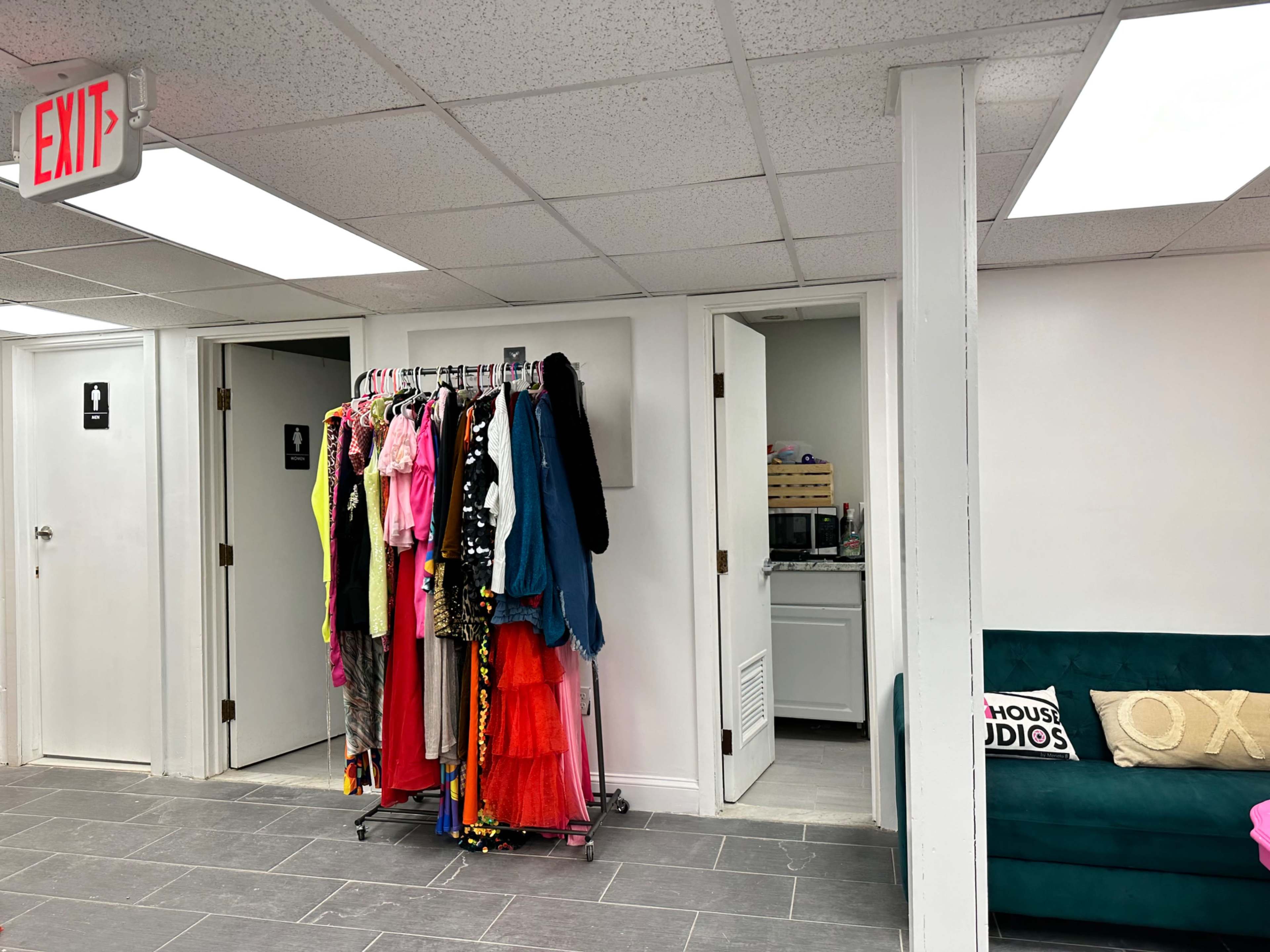 The image shows a brightly lit room featuring a clothing rack with various colorful outfits, a green couch, and signs for restrooms on the walls.