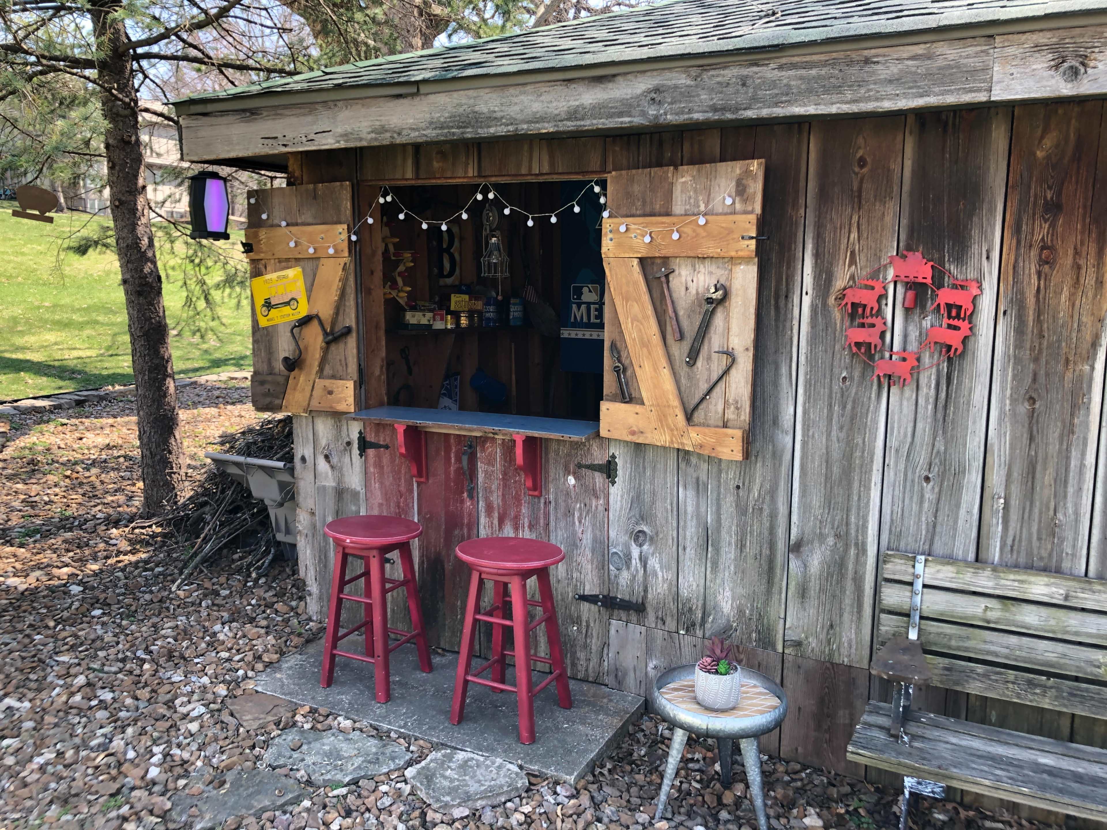 A wooden shed features a service window with open shutters and two red stools in front, set in a gravel area surrounded by greenery.