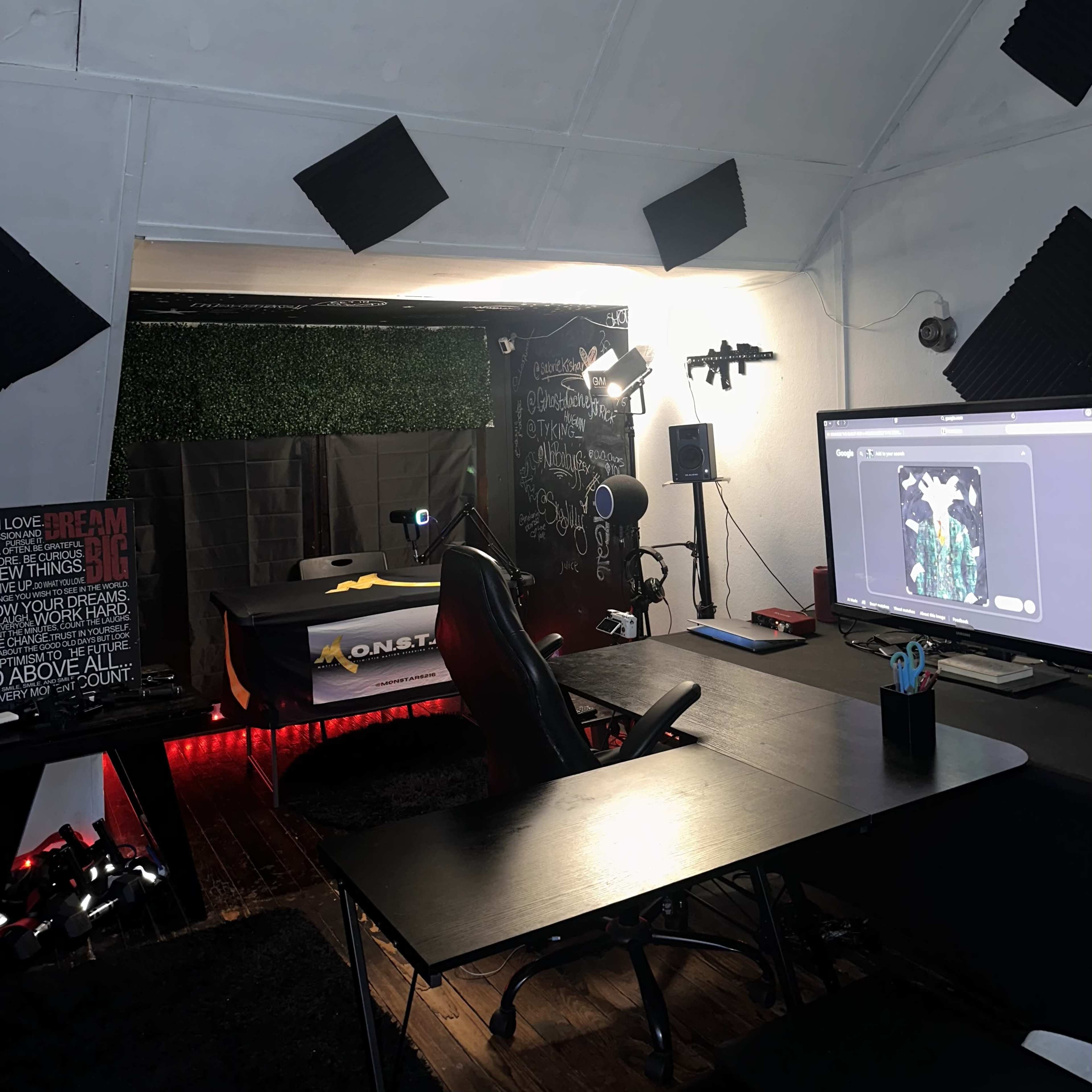 The image shows a small recording studio with a black backdrop, a large monitor displaying an image, and various equipment and soundproofing panels lining the walls.
