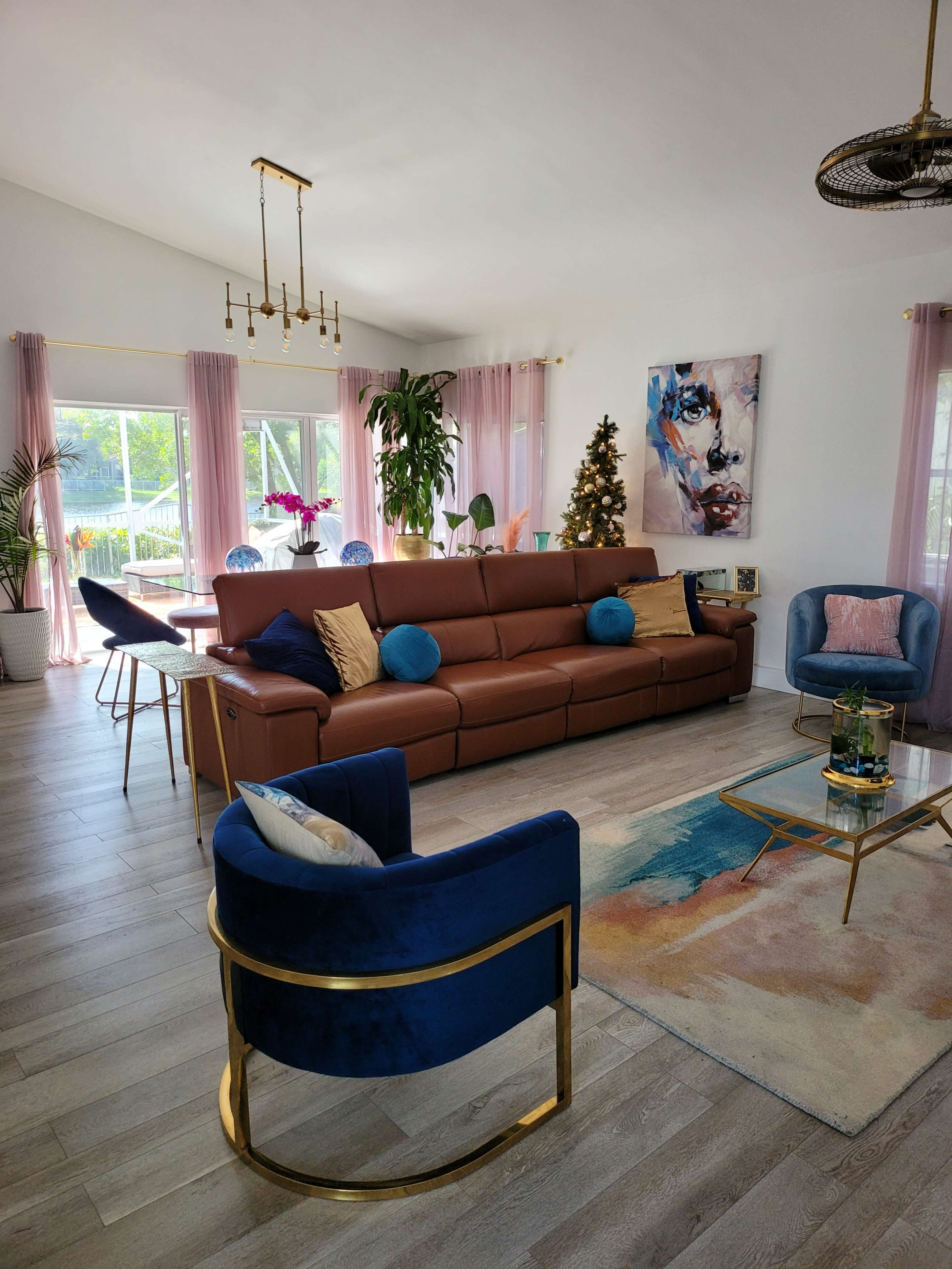 A modern living room with a brown sectional sofa, two blue accent chairs, a glass coffee table, and large windows displaying greenery outside.