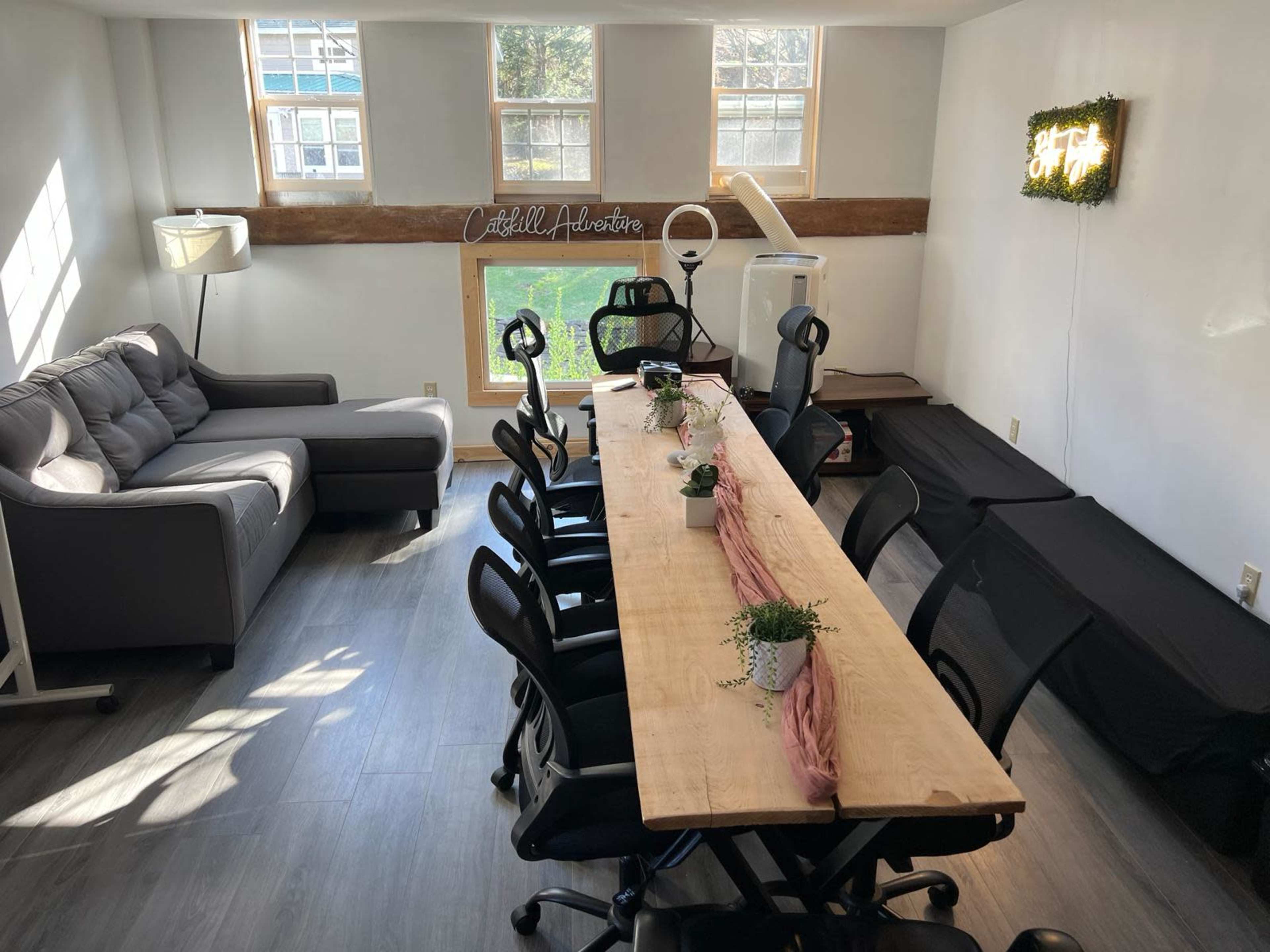 A well-lit meeting room with a long wooden table surrounded by black office chairs, a sofa in the corner, and a window that offers a view of greenery outside.