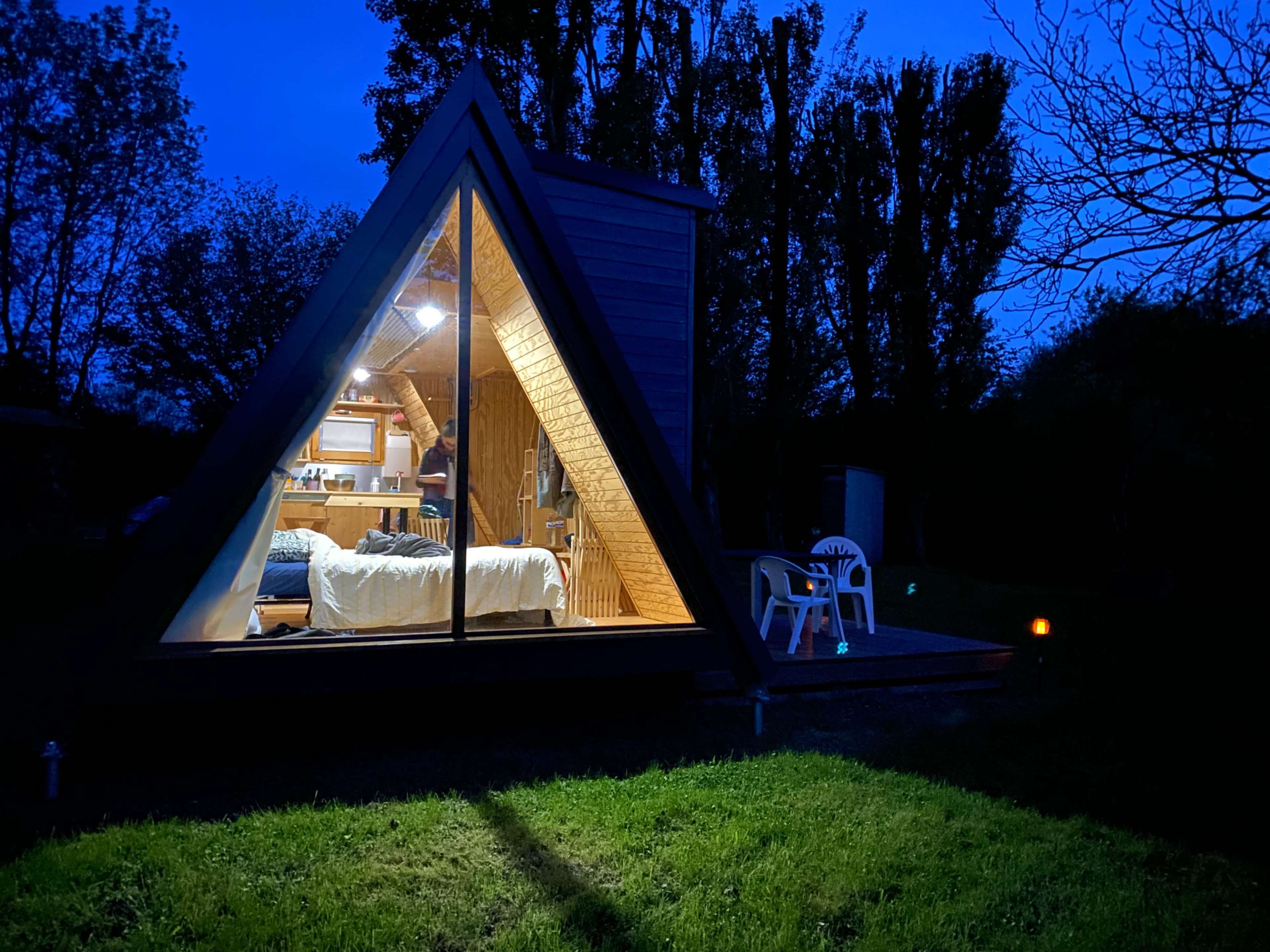 A triangular wooden cabin with a large glass window is illuminated at night, showing a cozy interior and surrounded by a grassy area.