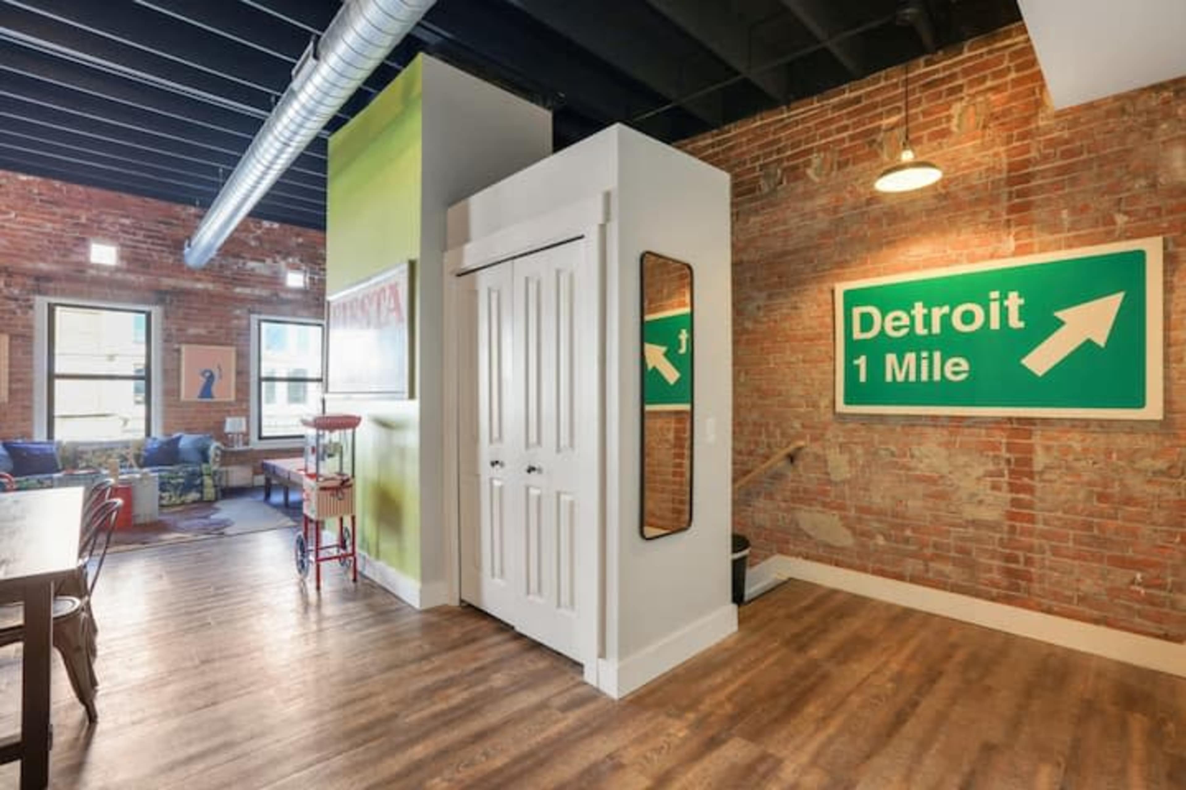 A contemporary interior space featuring exposed brick walls, hardwood flooring, and a decorative sign indicating the distance to Detroit.
