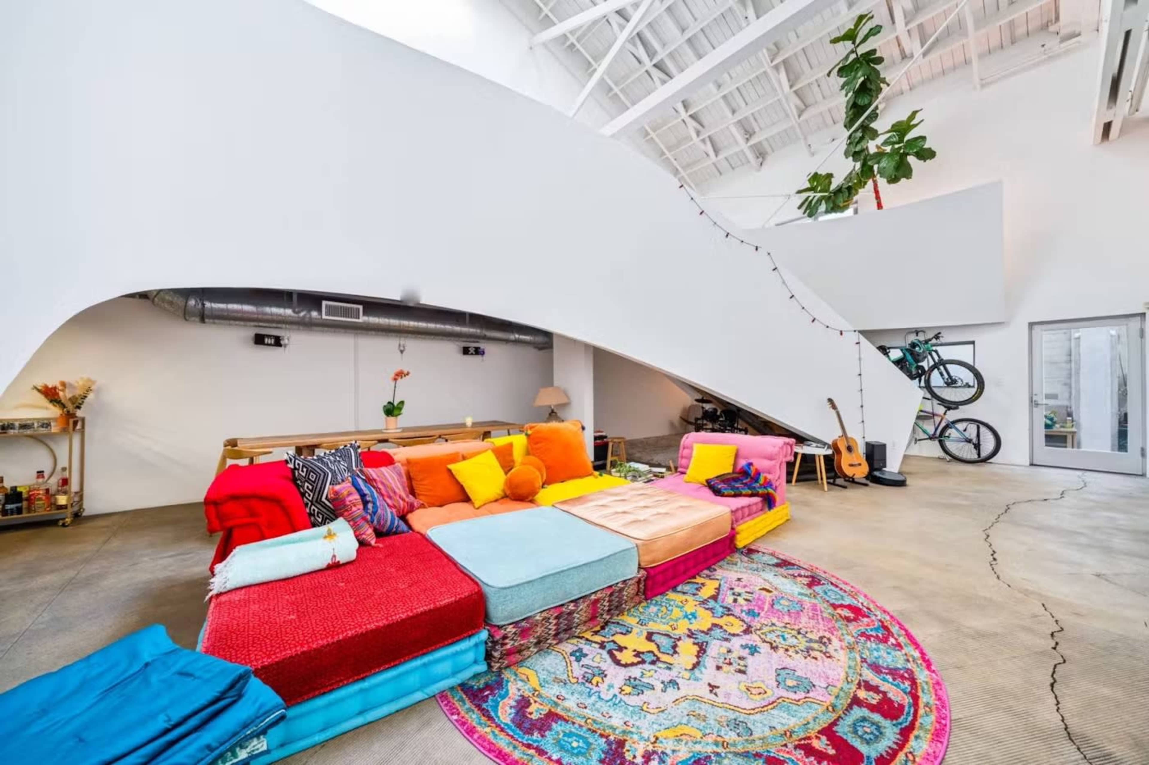A brightly colored lounge area with a large, multi-colored sectional sofa, a circular patterned rug, and potted plants, all within a spacious, modern room featuring high ceilings.