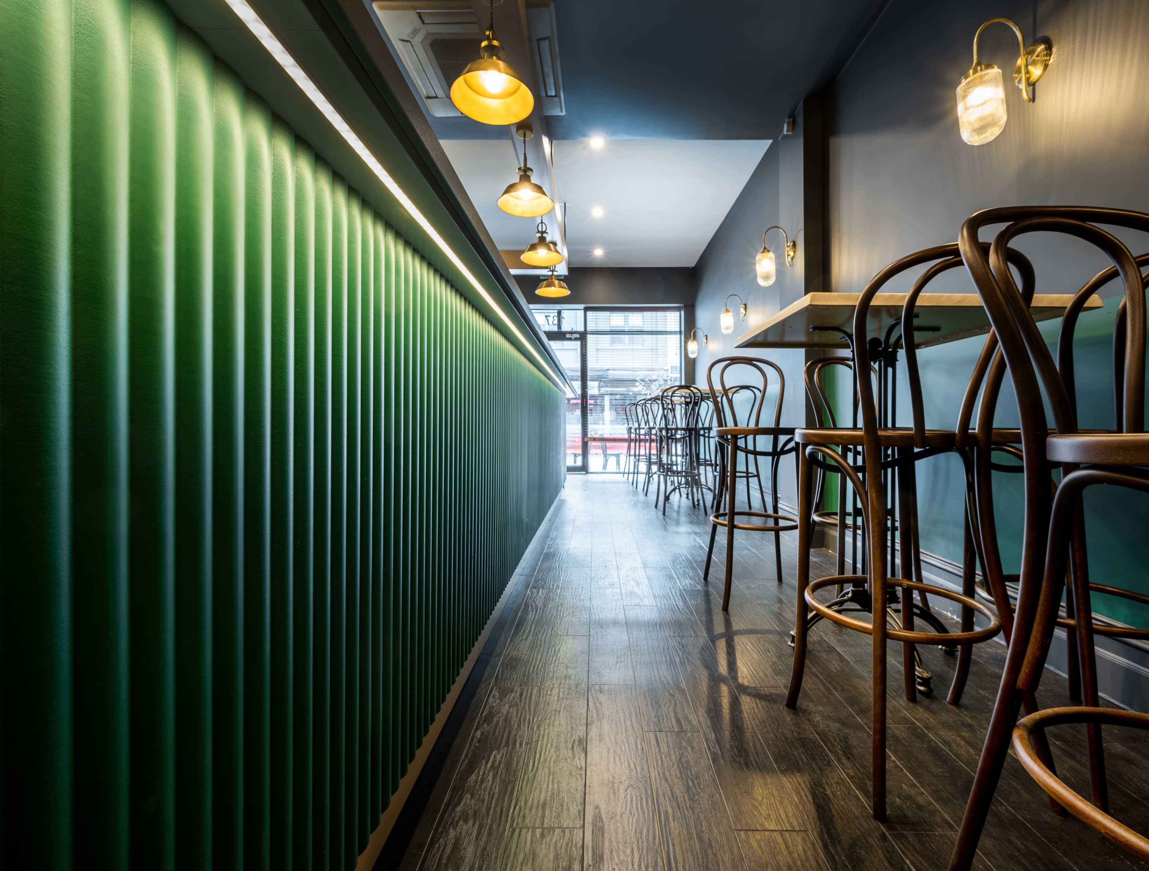 The image shows a modern café interior featuring green paneled walls, wooden flooring, and high stools arranged along a counter with pendant lighting.