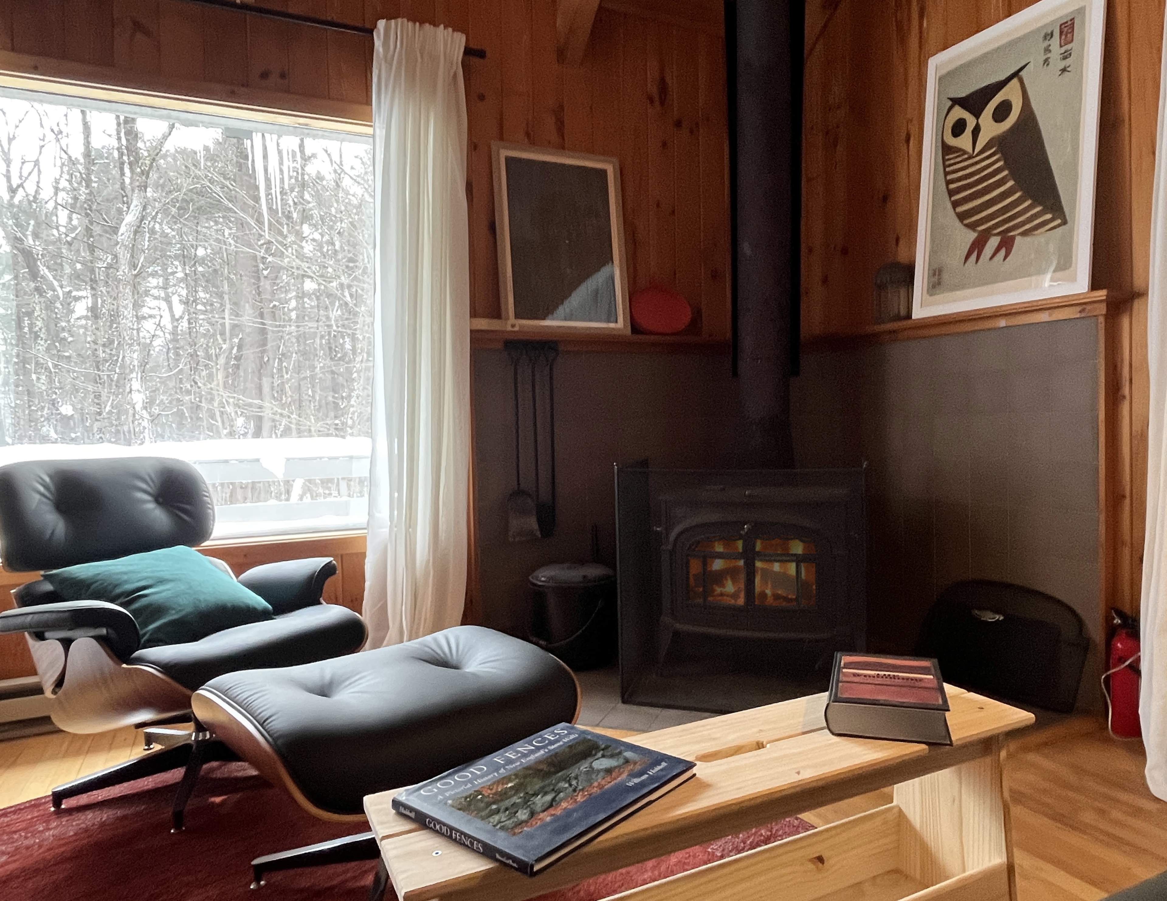 The image features a cozy wooden interior with a fireplace, a black lounge chair and ottoman, and a coffee table topped with books, all overlooking a snowy landscape outside the window.