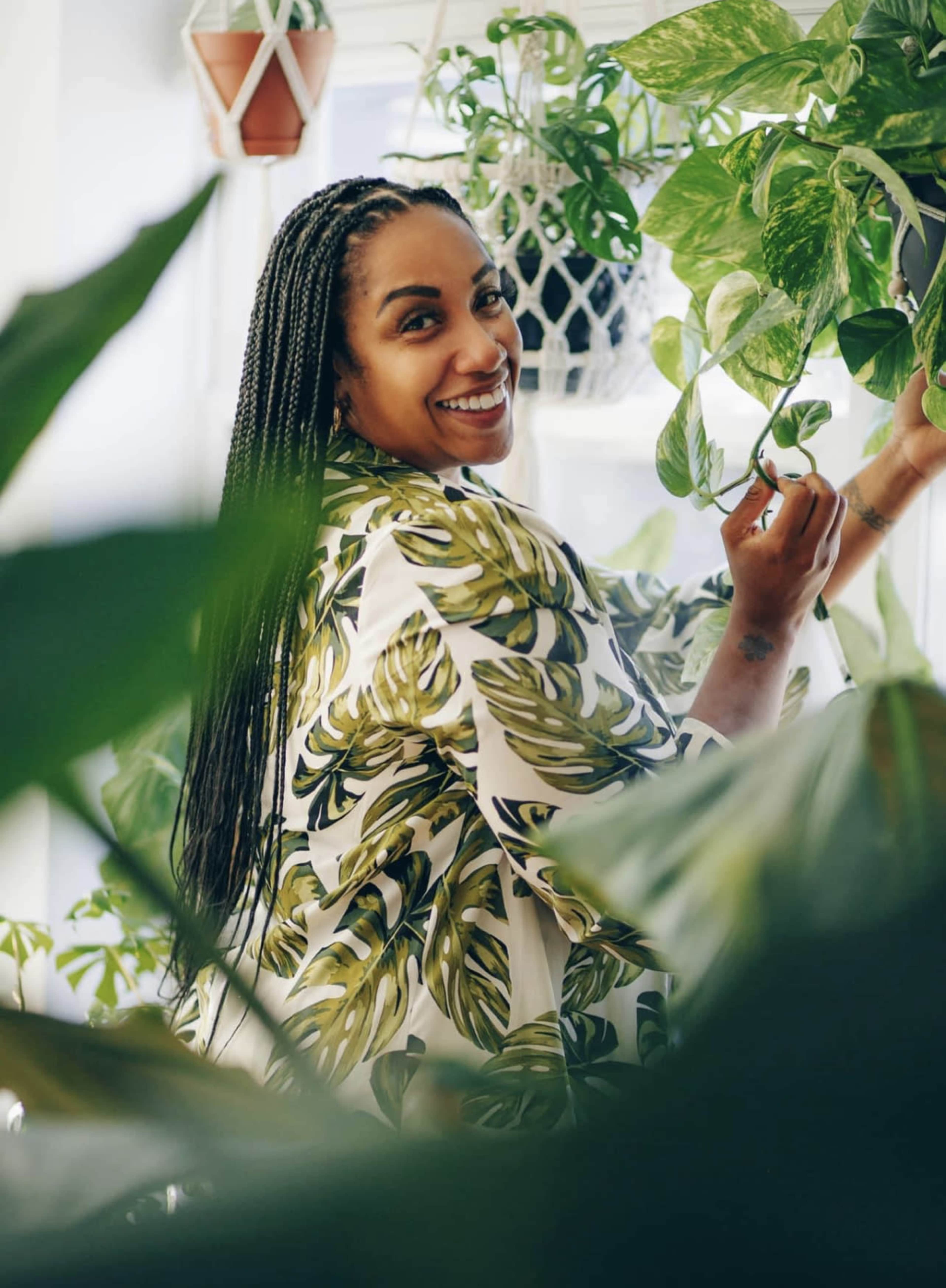 A woman with braided hair is smiling while tending to potted plants in a bright indoor setting.