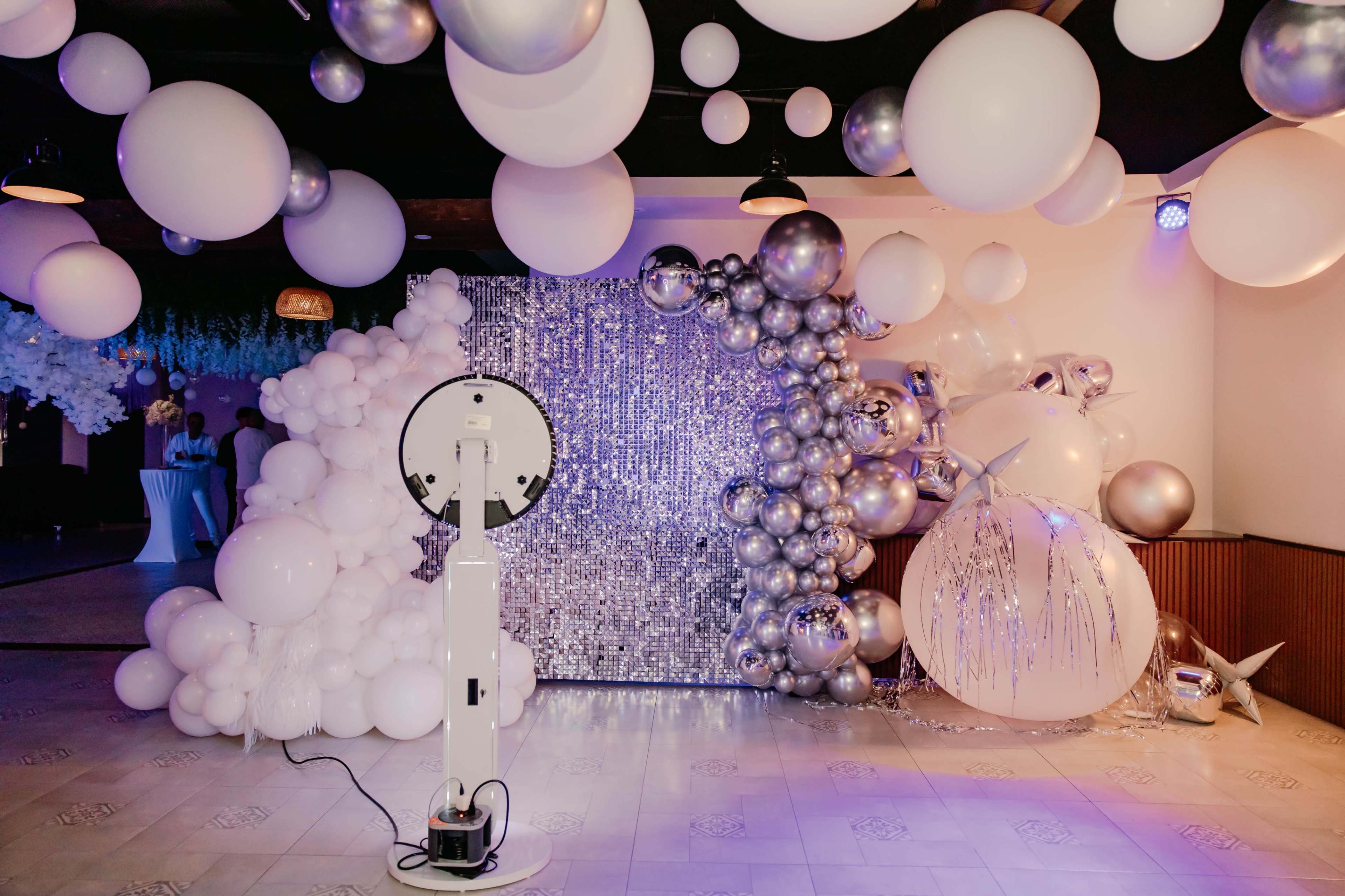The image shows a decorated party space featuring a backdrop of silver balloons and a sequined wall, with a photo booth setup in front.