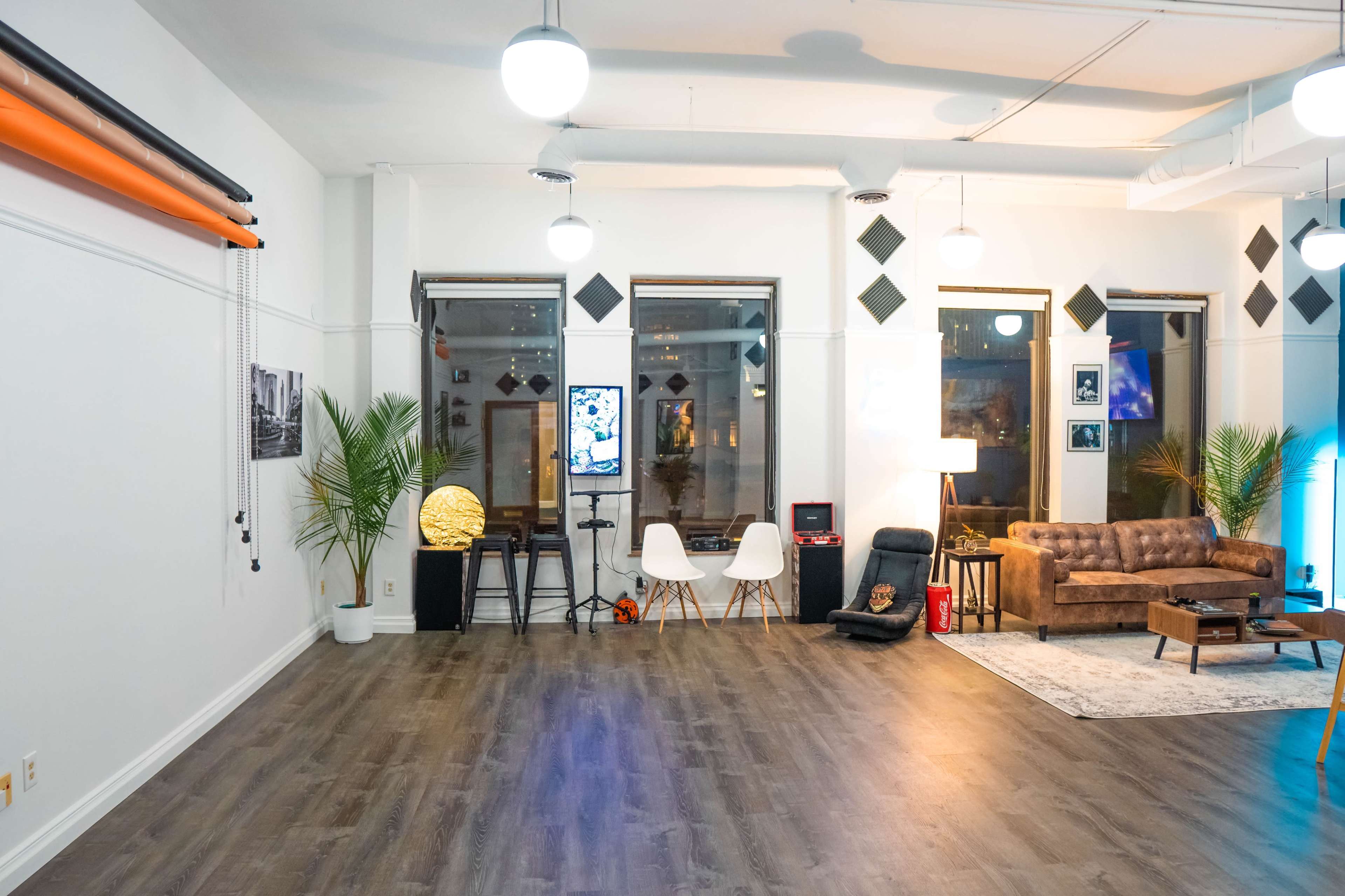 A spacious room with wooden flooring, featuring a brown leather sofa, a small table, and decorative plants alongside a large window displaying city lights.