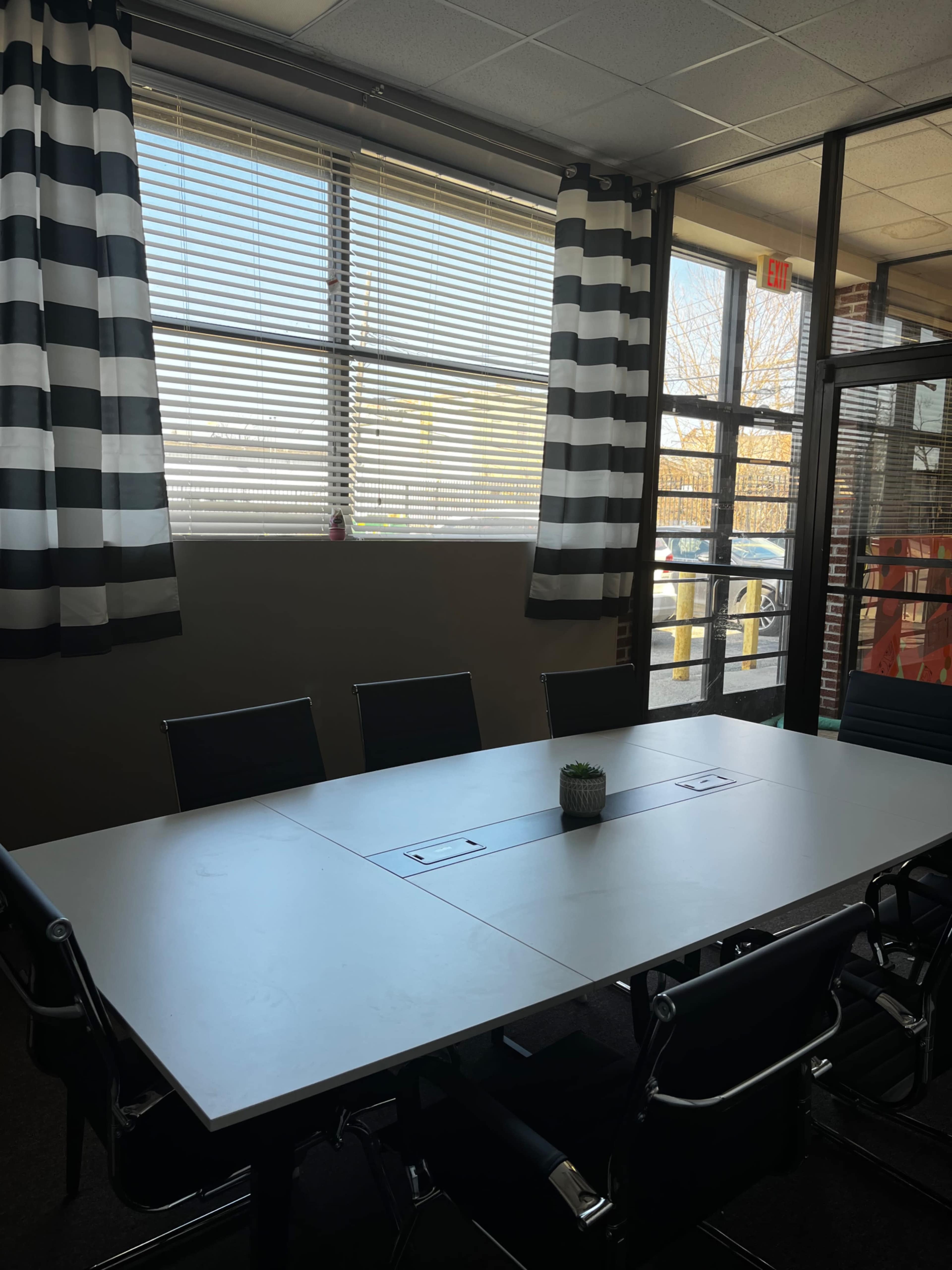 A conference room with a large table surrounded by black chairs, sunlight streaming in through large windows with striped curtains.
