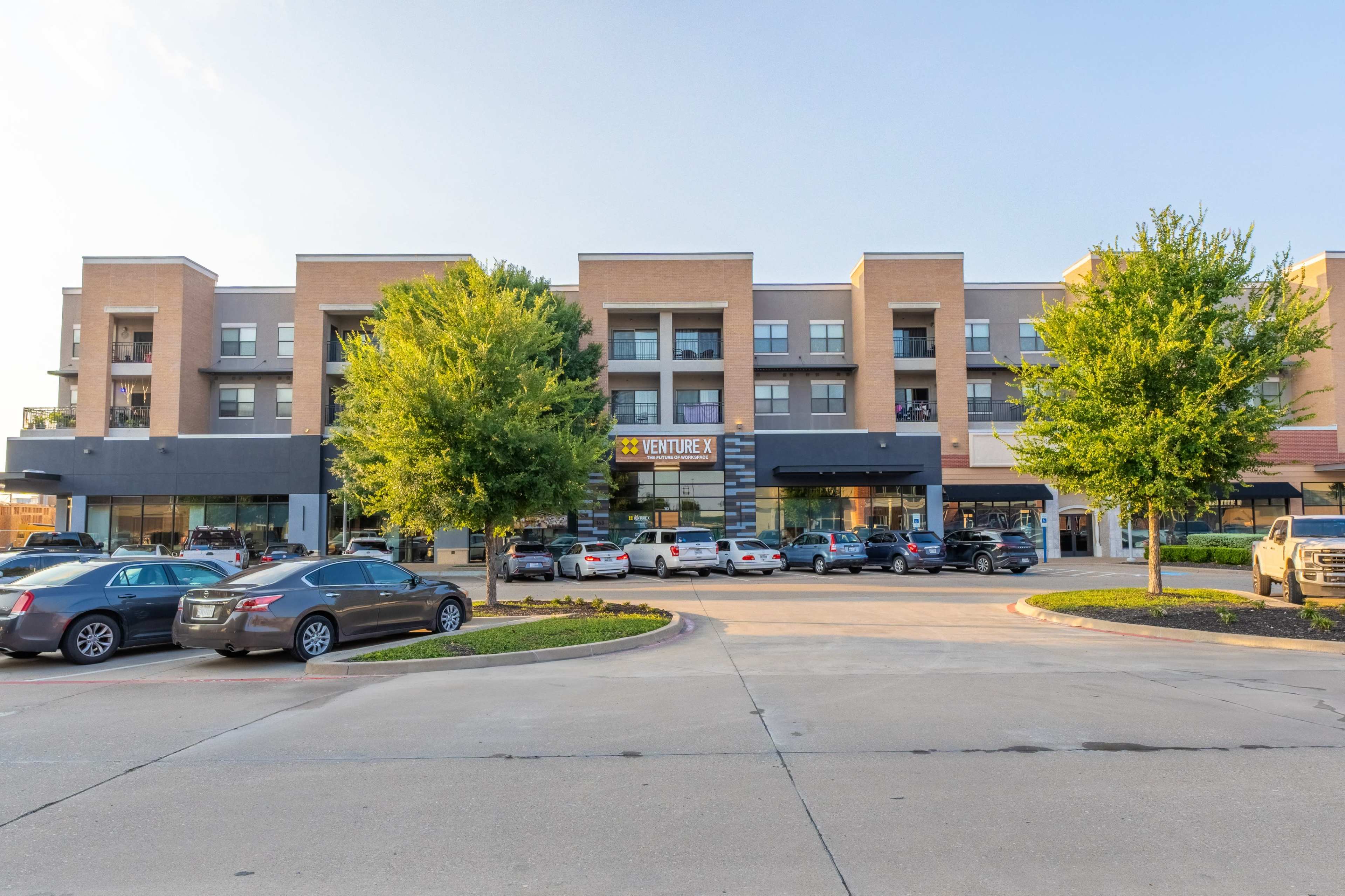 A multi-story brick commercial building features shops on the ground floor and residential units above, surrounded by parked cars and trees.