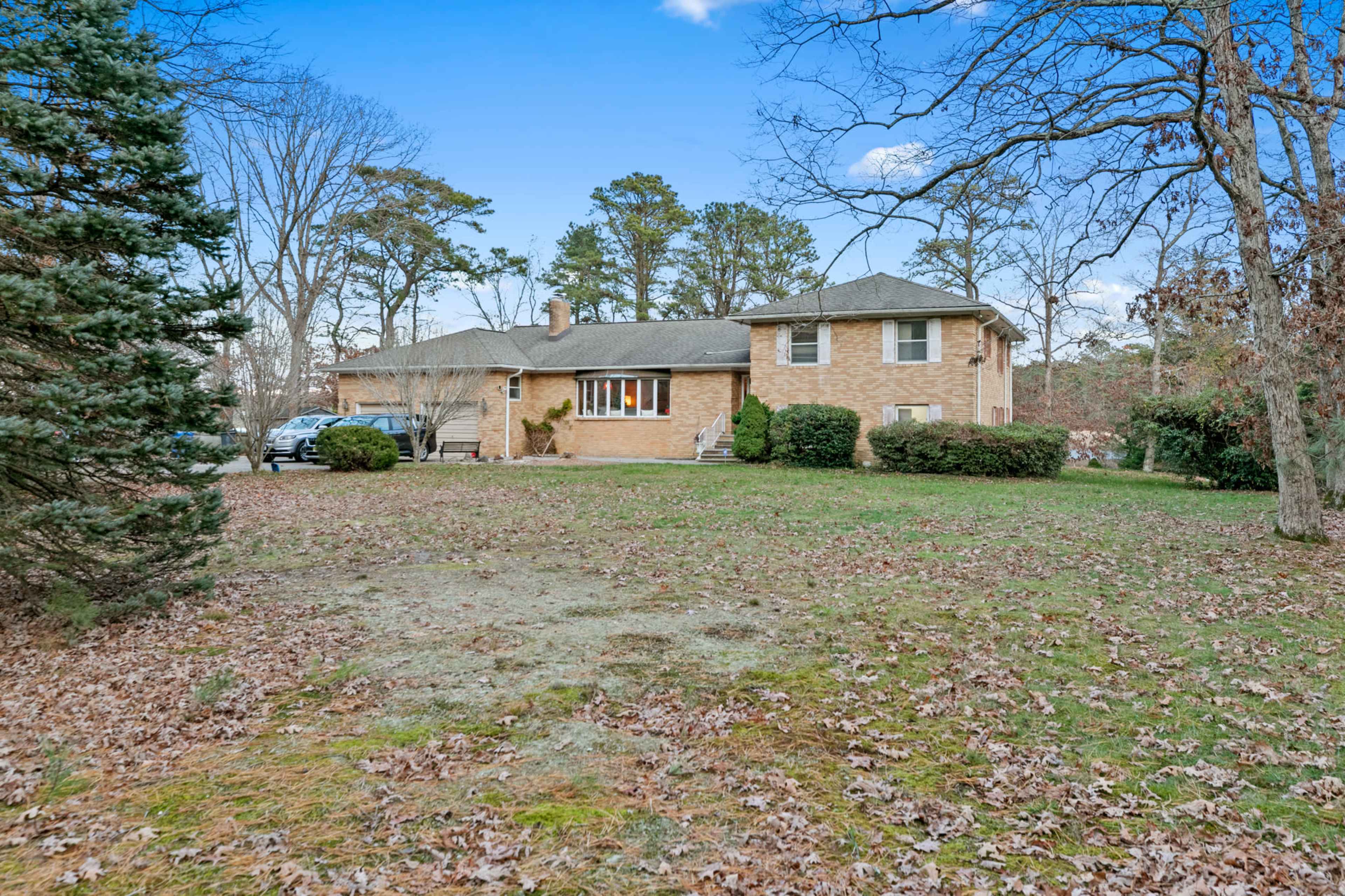 A brick house with a front porch is set in a yard featuring fallen leaves and trees in the background.