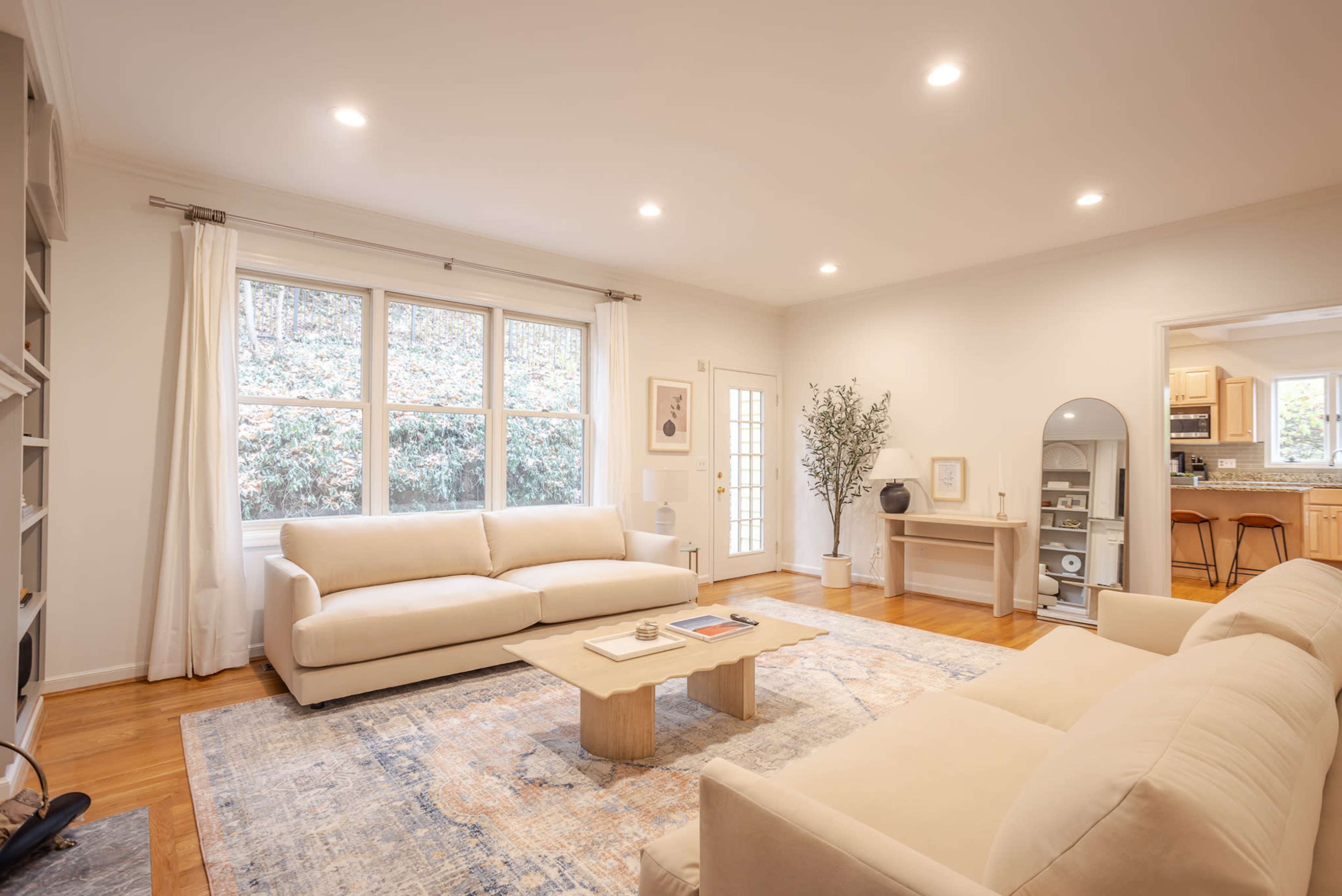 The living room features two light-colored sofas facing a central coffee table, with large windows providing natural light and a view of outdoor greenery.