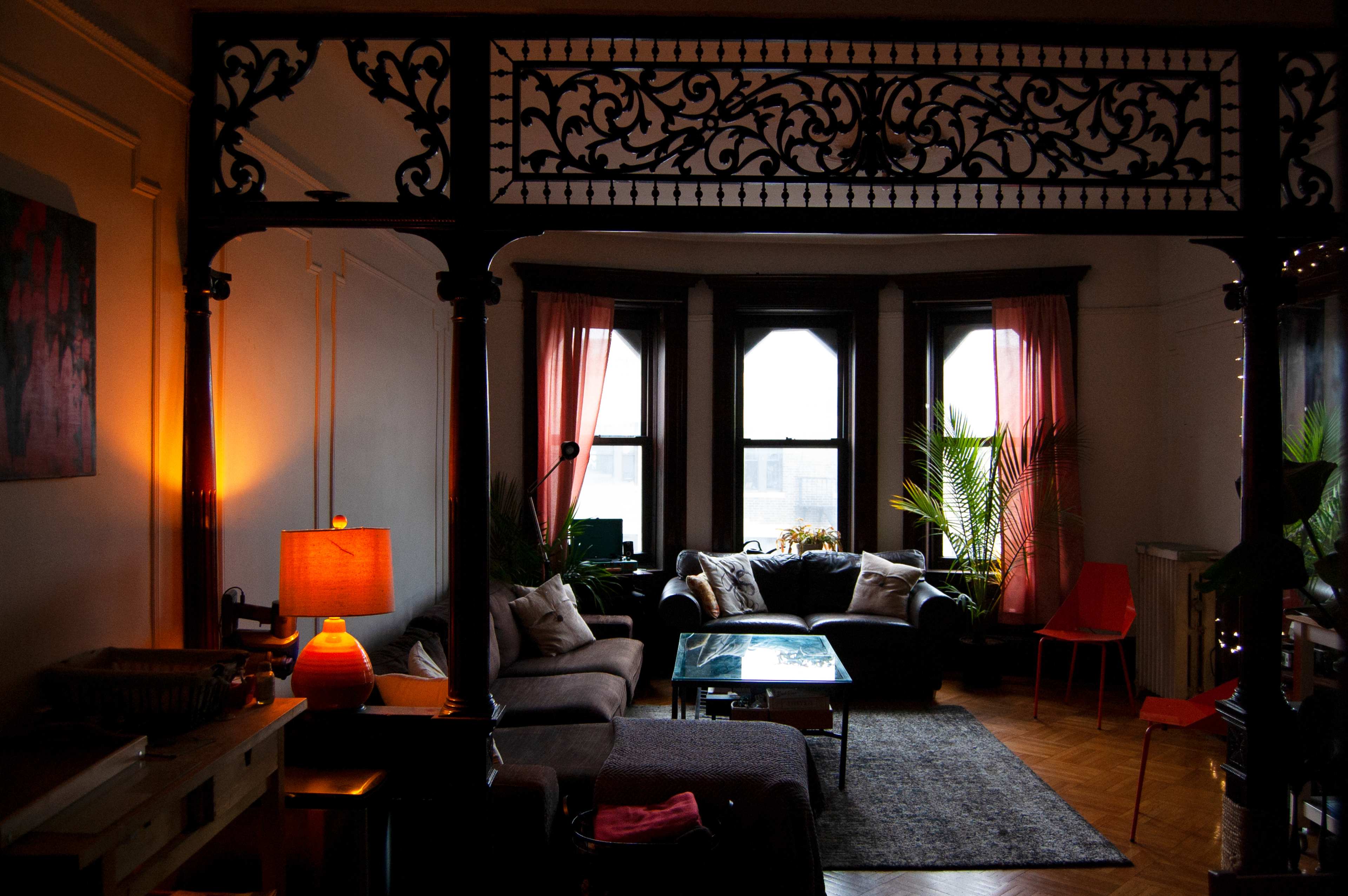 The scene features a living room with large windows draped in pink curtains, a sectional sofa, a glass coffee table, and decorative plants, all framed by an ornate architectural divider.