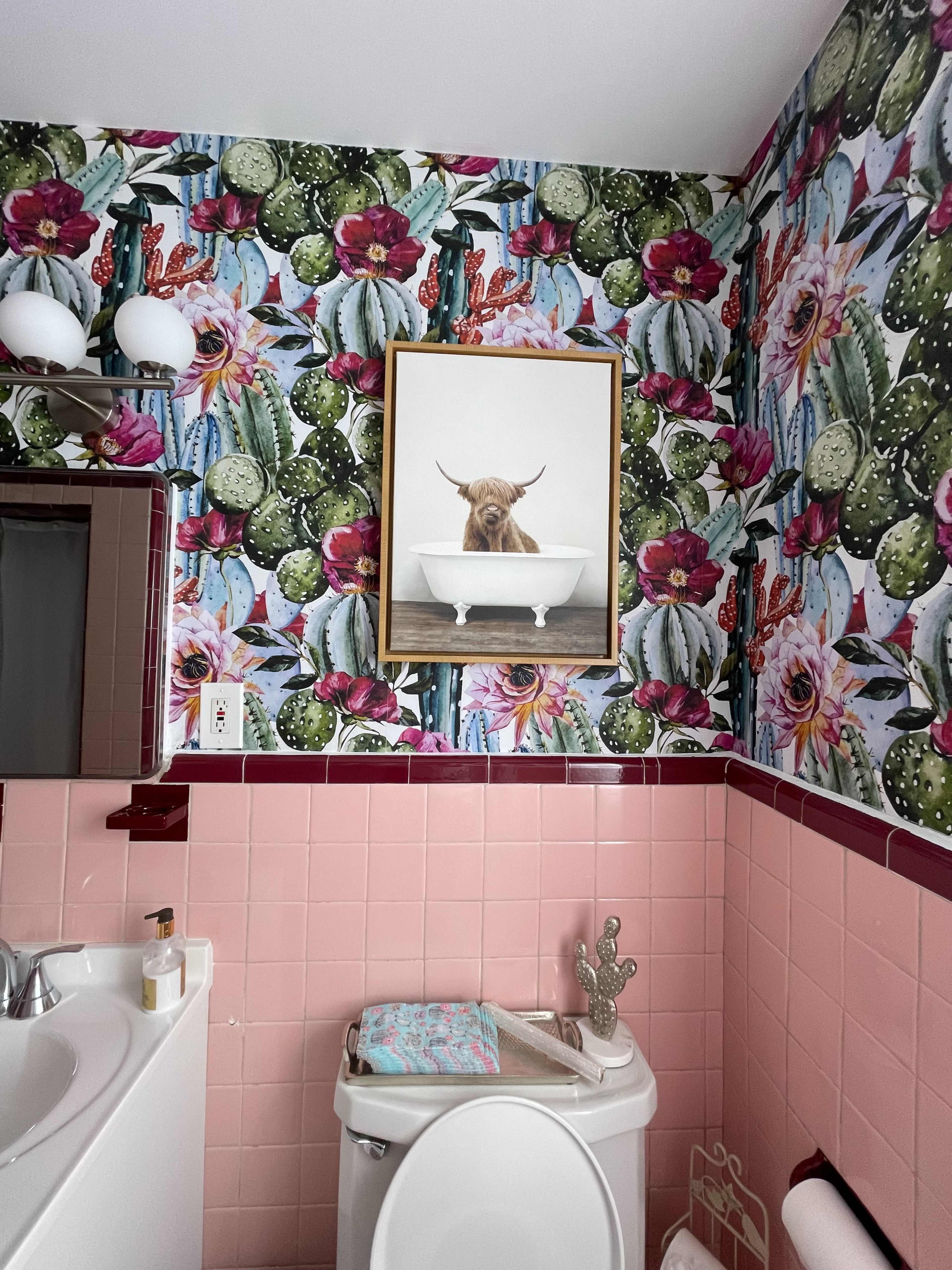 The bathroom features floral and cactus wallpaper, a framed picture of a cow above a white bathtub, and light pink tiled walls.