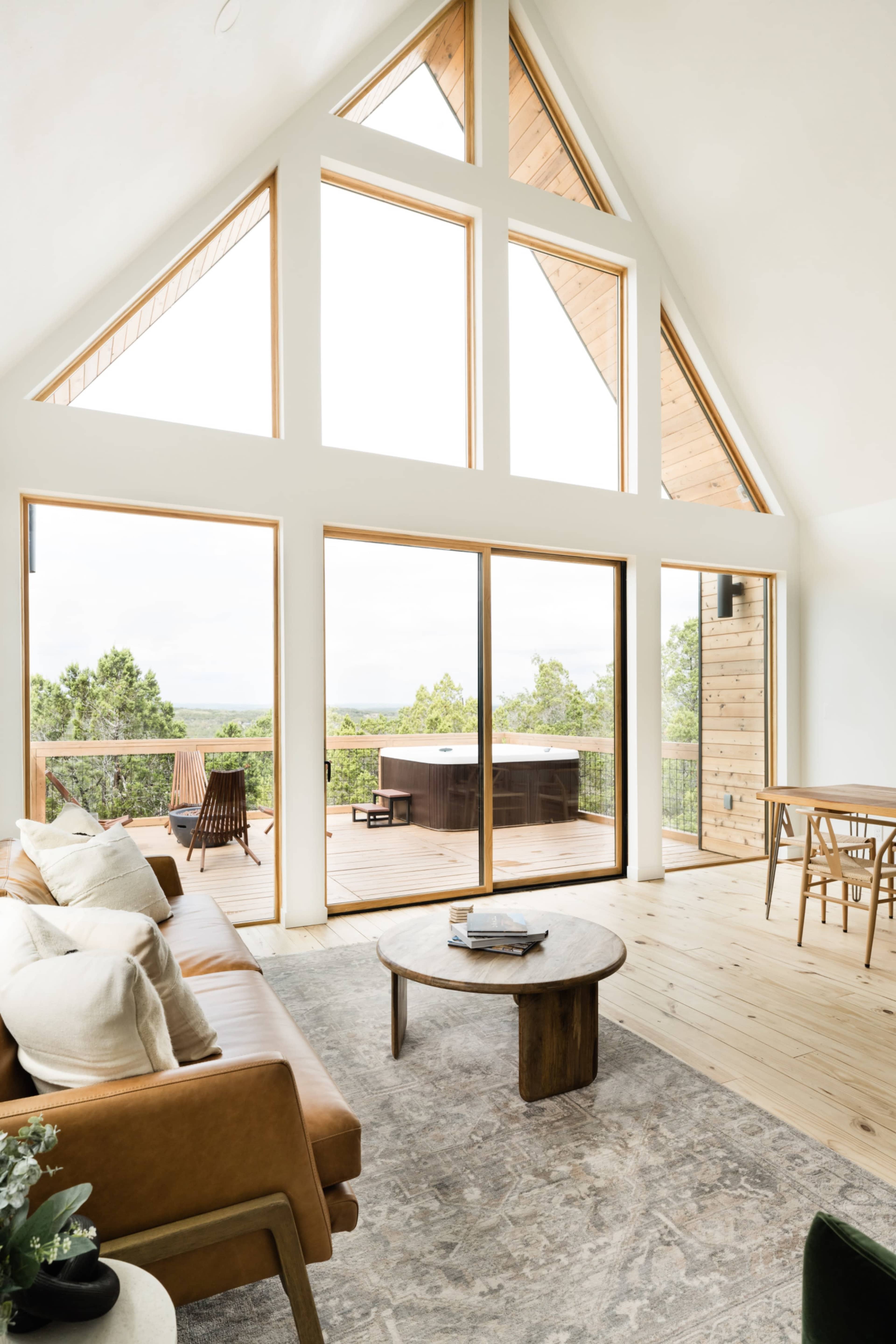 The interior of a modern, triangular-roofed cabin features large windows, a cozy seating area, and a deck with a hot tub overlooking greenery.