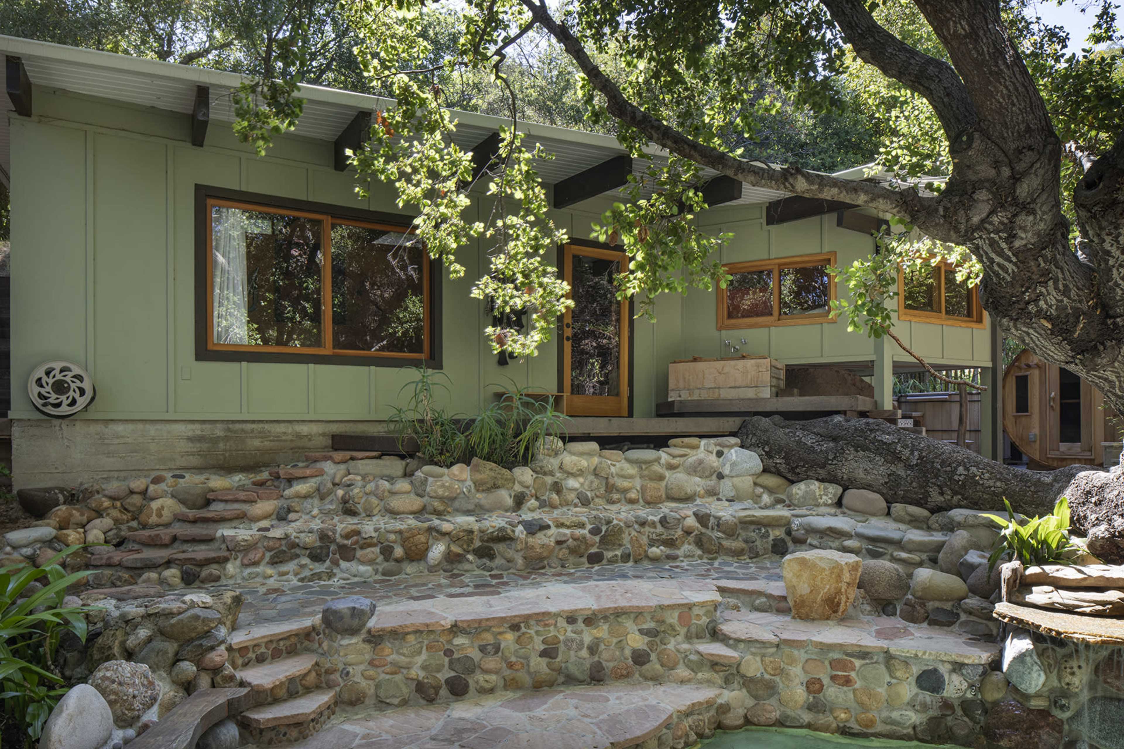 A green house with large windows is surrounded by rocks and greenery, featuring a stone pathway and a patio area.