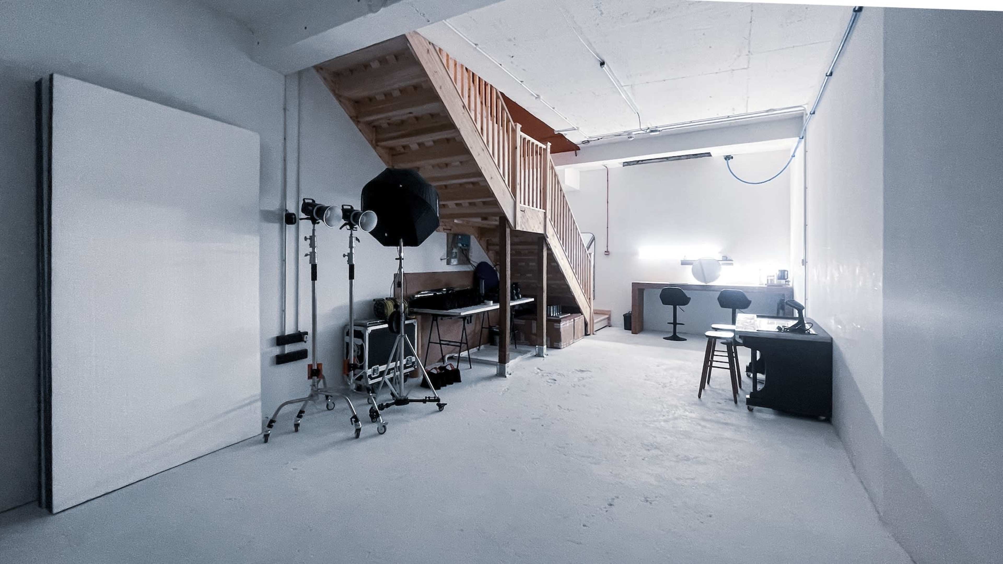 The image shows a spacious room with a staircase on the left, a table and chairs in the background, and photography equipment set up near the center.