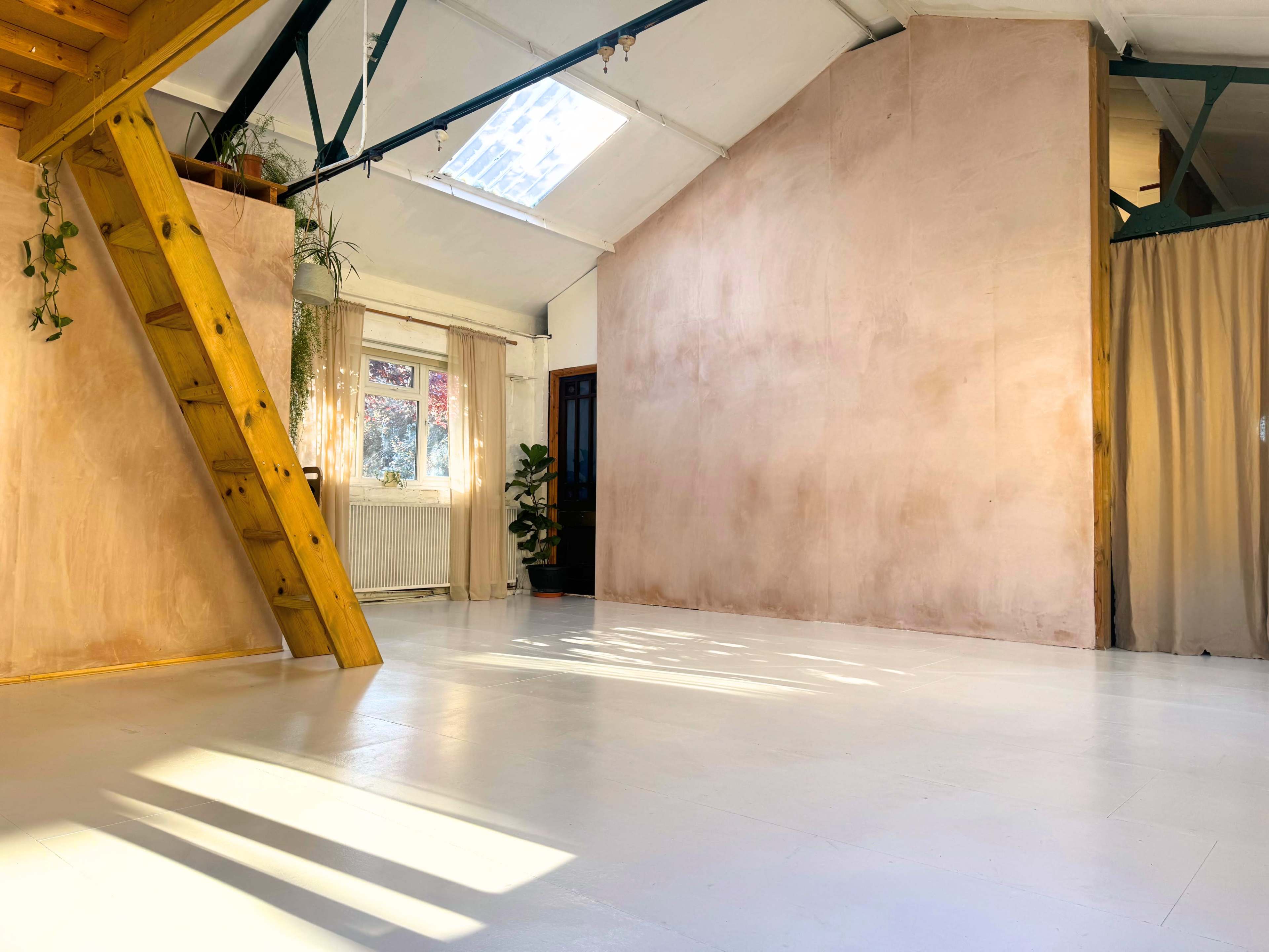 A spacious room with a wooden ladder, light-colored walls, and sunlight casting shadows on the white floor.