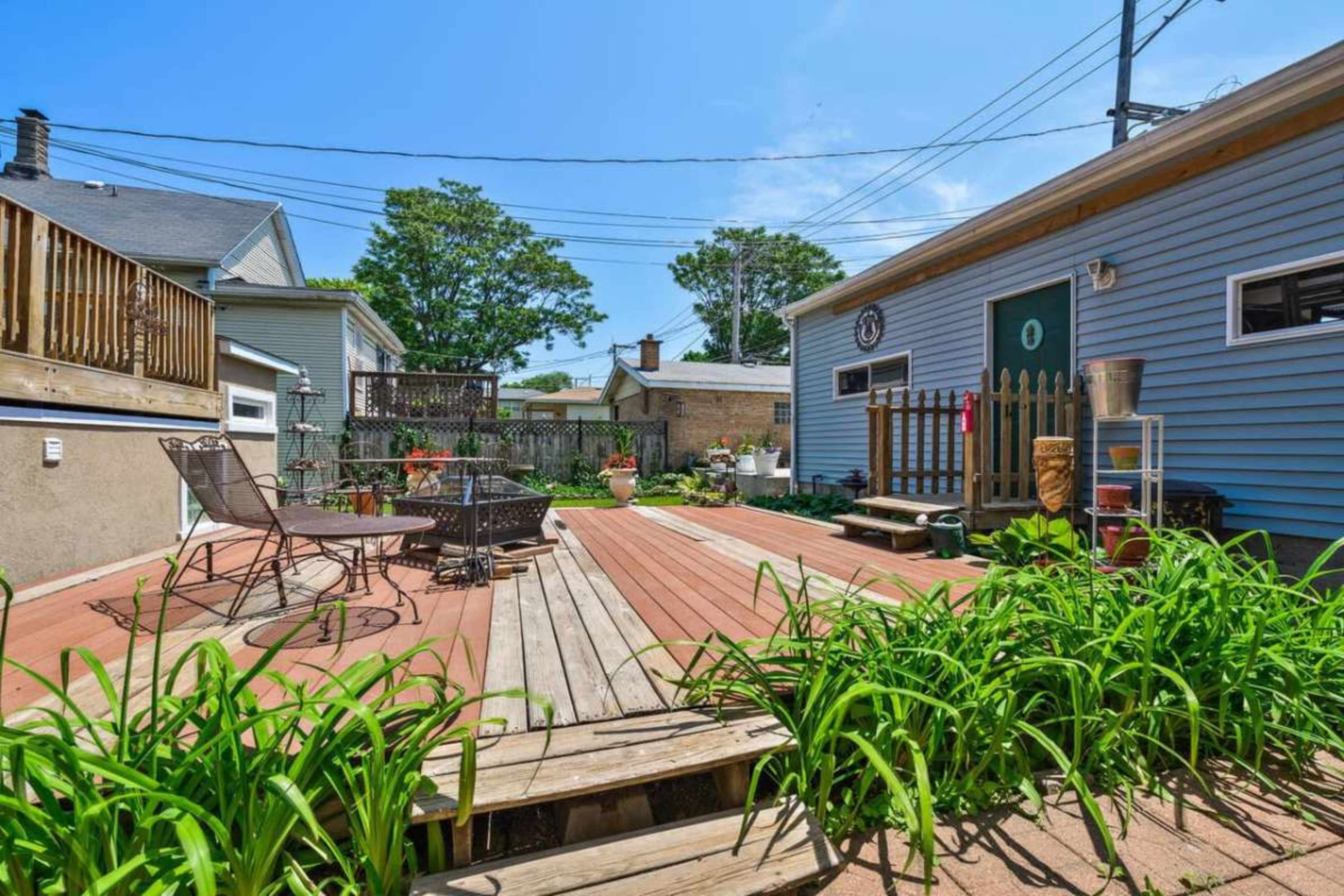 A wooden deck with potted plants is seen leading to a blue house with a door and a circular window, surrounded by greenery and power lines above.