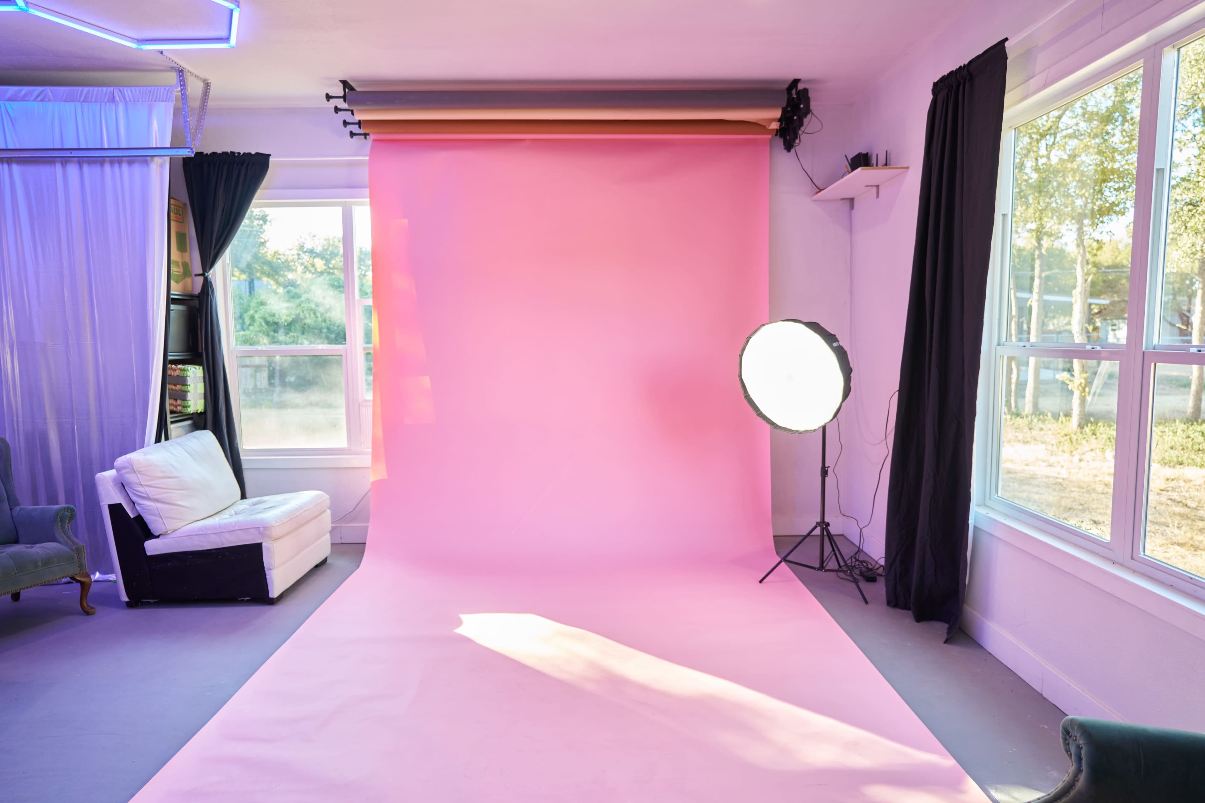 The image shows a photography studio with a pink backdrop, a large light source on a stand, and a white armchair nearby.