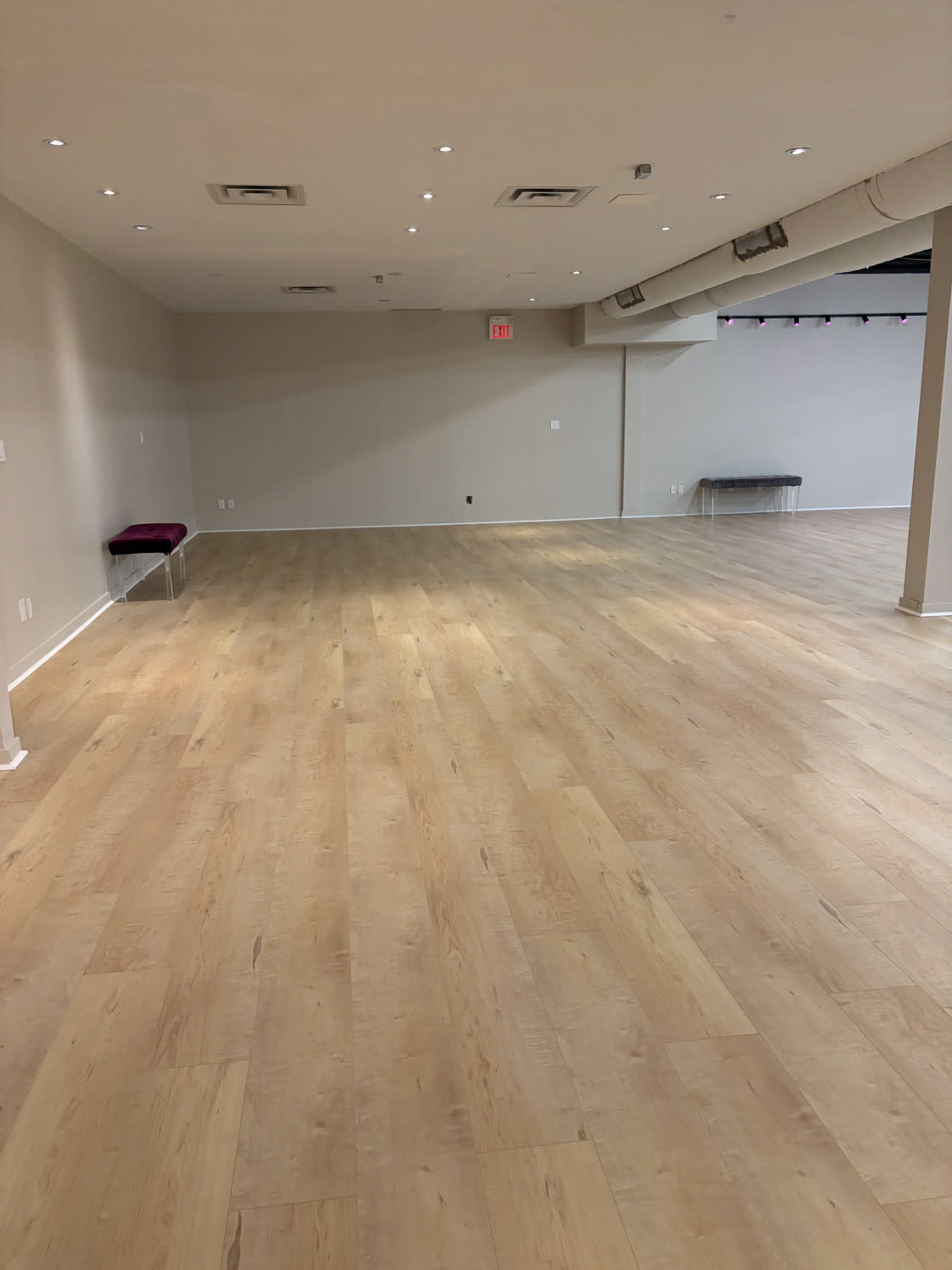 The image shows a large, empty space with wooden flooring and neutral-colored walls, featuring some benches along the sides.