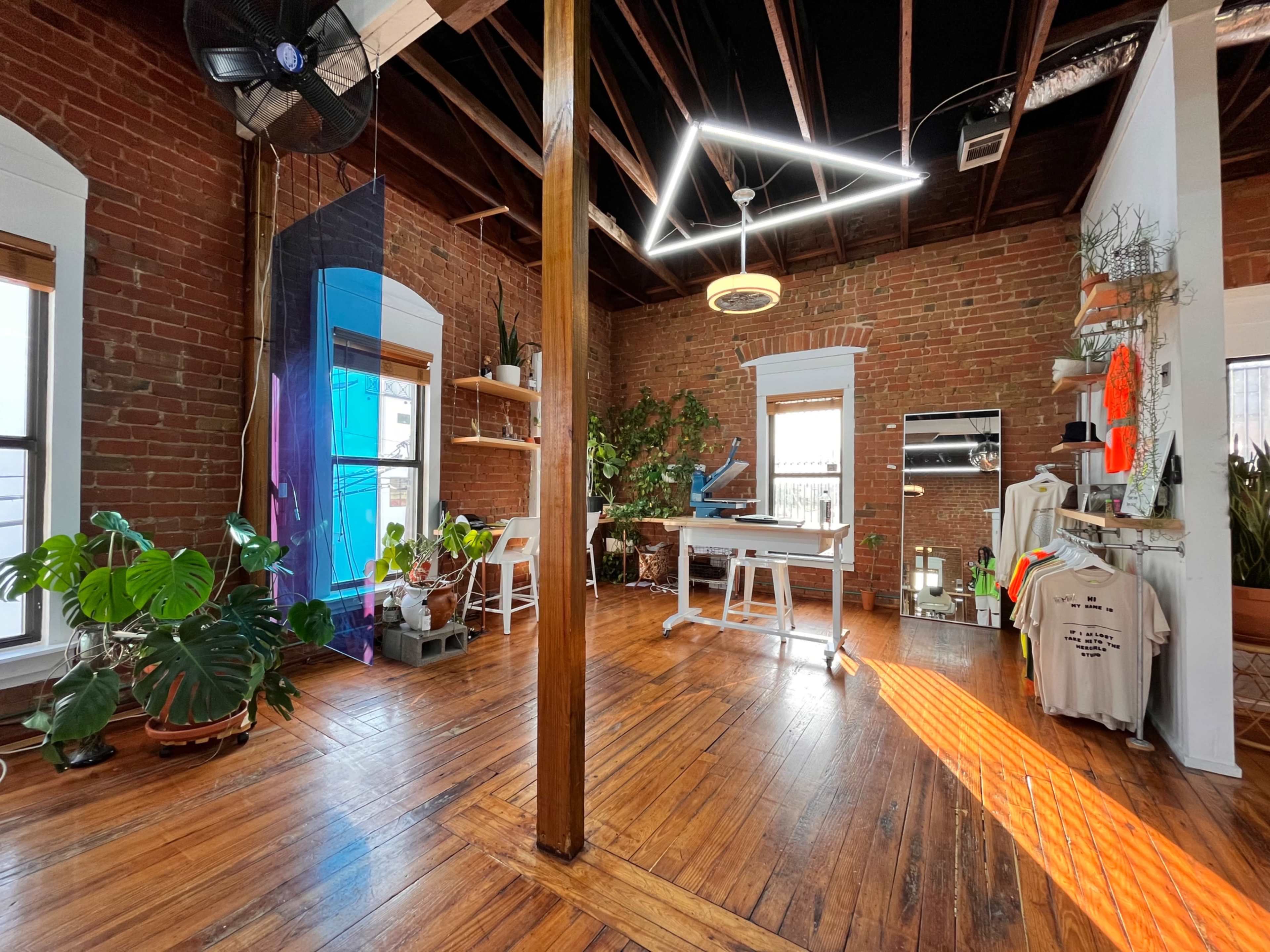 A spacious room with exposed brick walls, wooden floors, and various indoor plants, featuring a large work table, colorful decor items, and bright natural light streaming through the windows.
