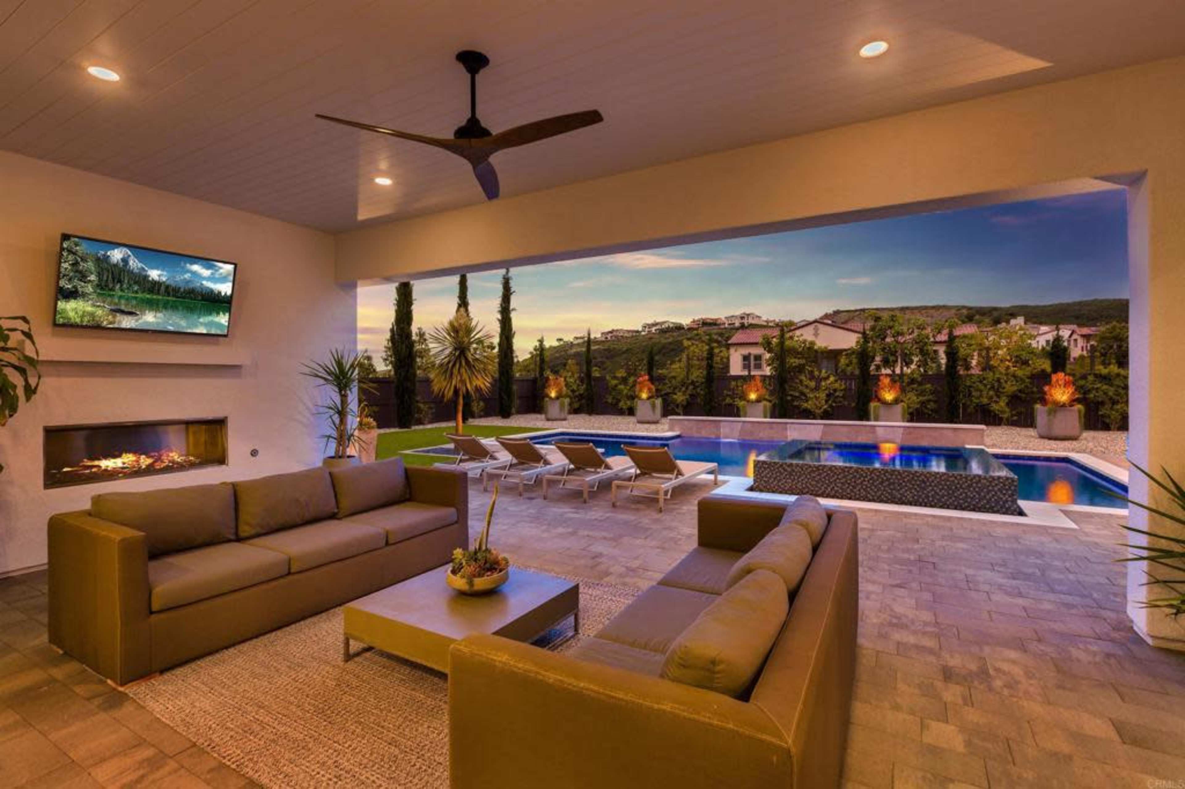 The image shows a modern outdoor lounge area with seating, a television, and a view of a swimming pool surrounded by landscaped gardens.