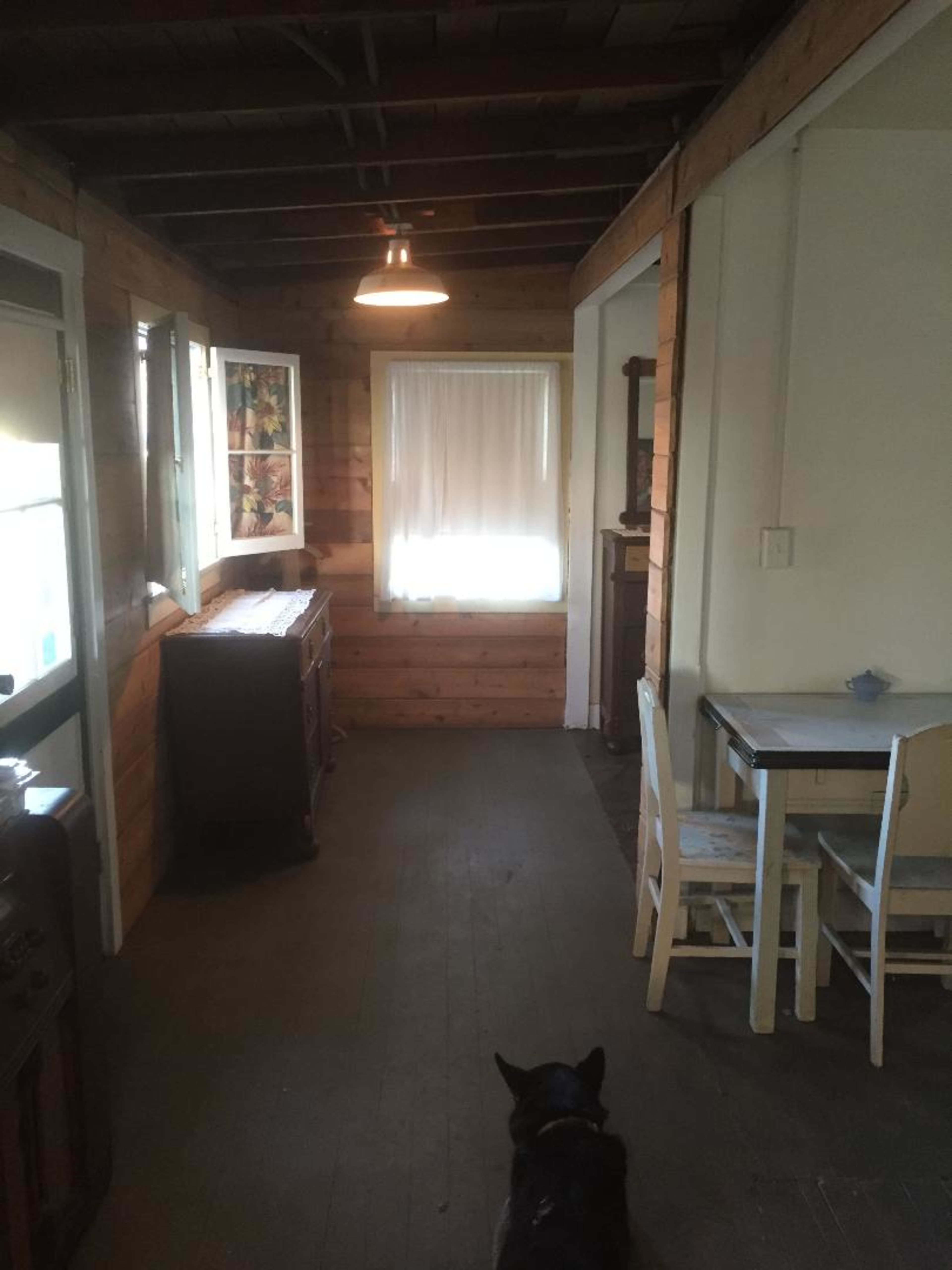 The image shows a hallway in a wooden cabin with a dining table and chairs on one side, a sideboard against the wall, and a dog standing on a wooden floor.