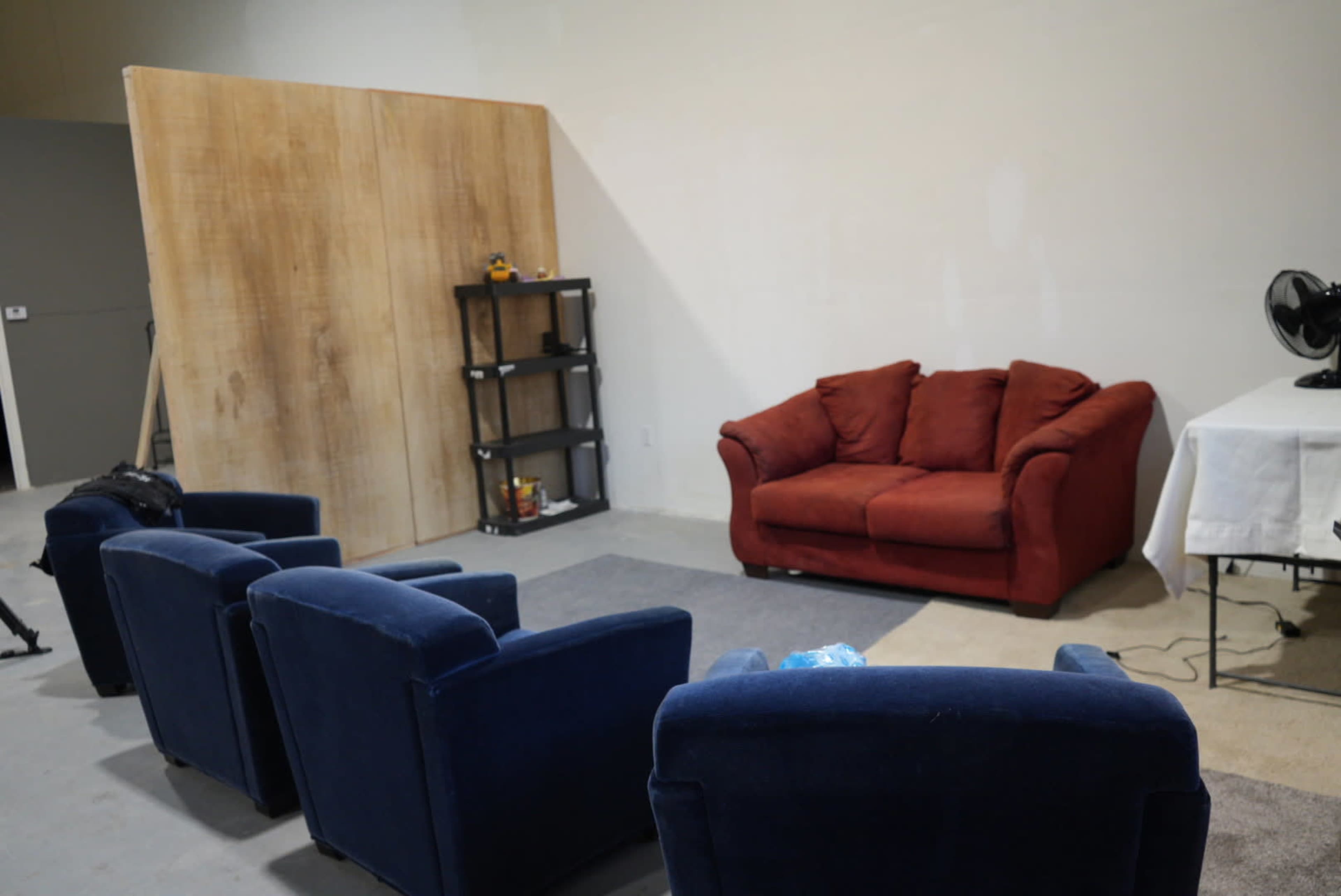 The image shows a sparse interior space featuring a red couch, several blue armchairs, a small black shelf, and a table covered with a white tablecloth against a plain wall.