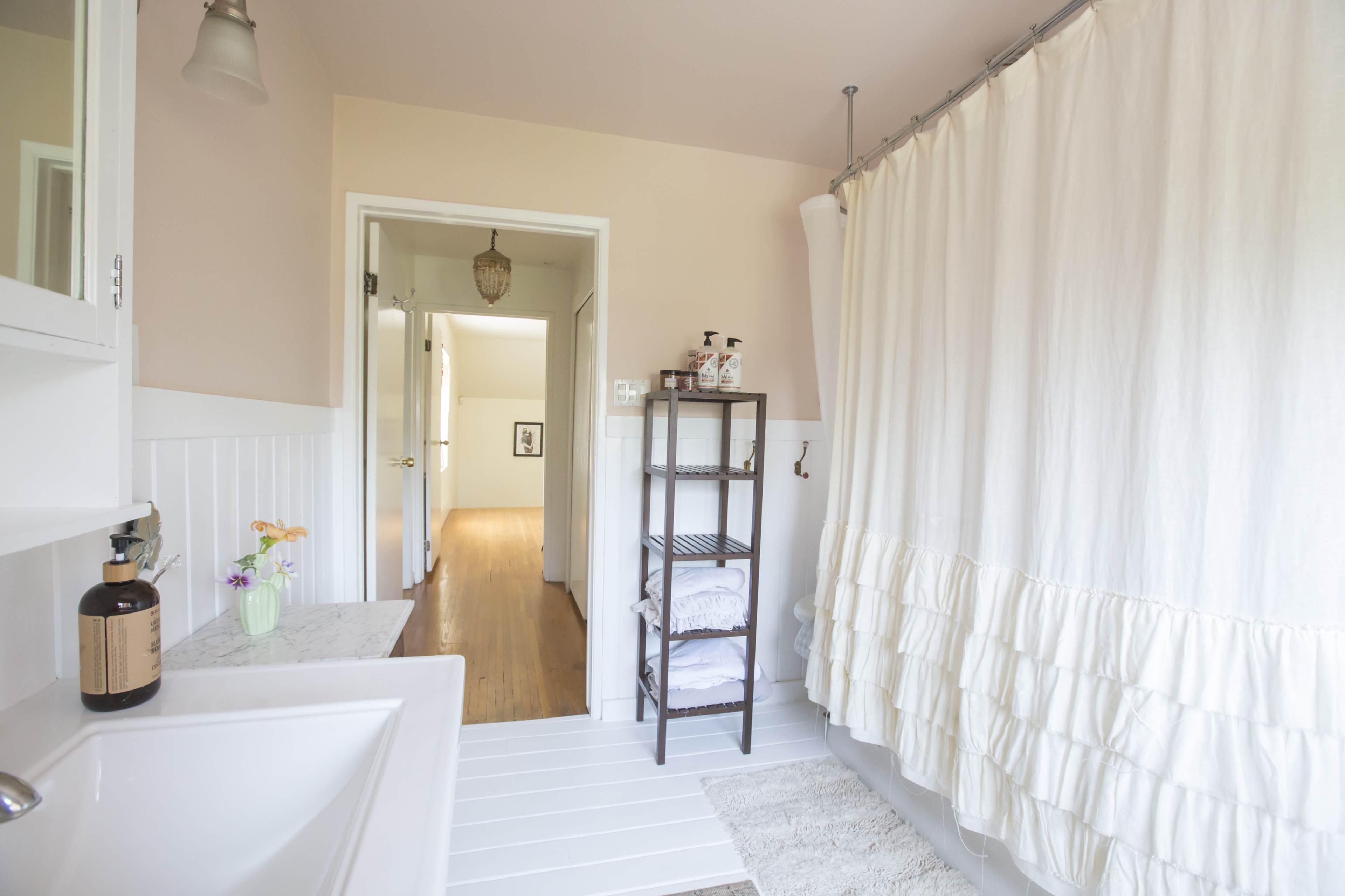 The image shows a bright bathroom with a white shower curtain, a marble countertop with a soap dispenser, and a shelf holding folded towels.