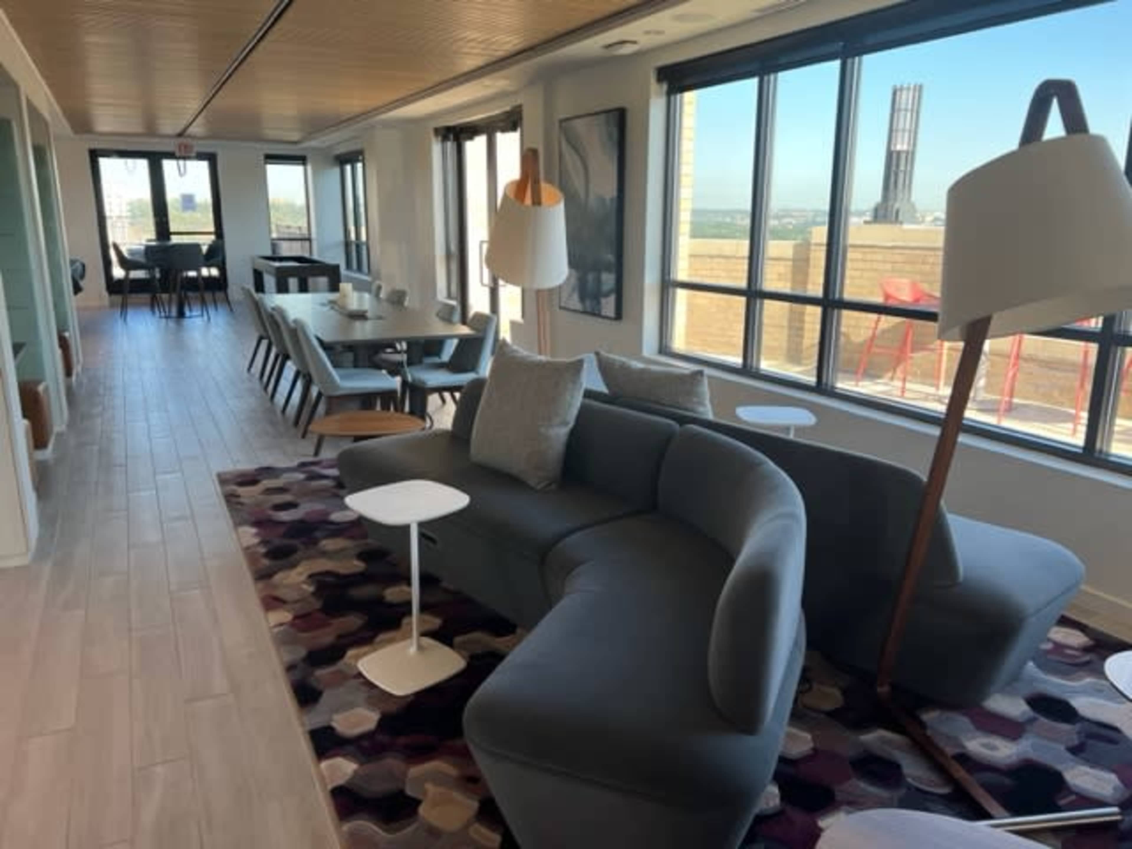 A modern lounge area featuring a curved gray sofa, decorative cushions, a standing lamp, and a small side table, with large windows providing views outside.