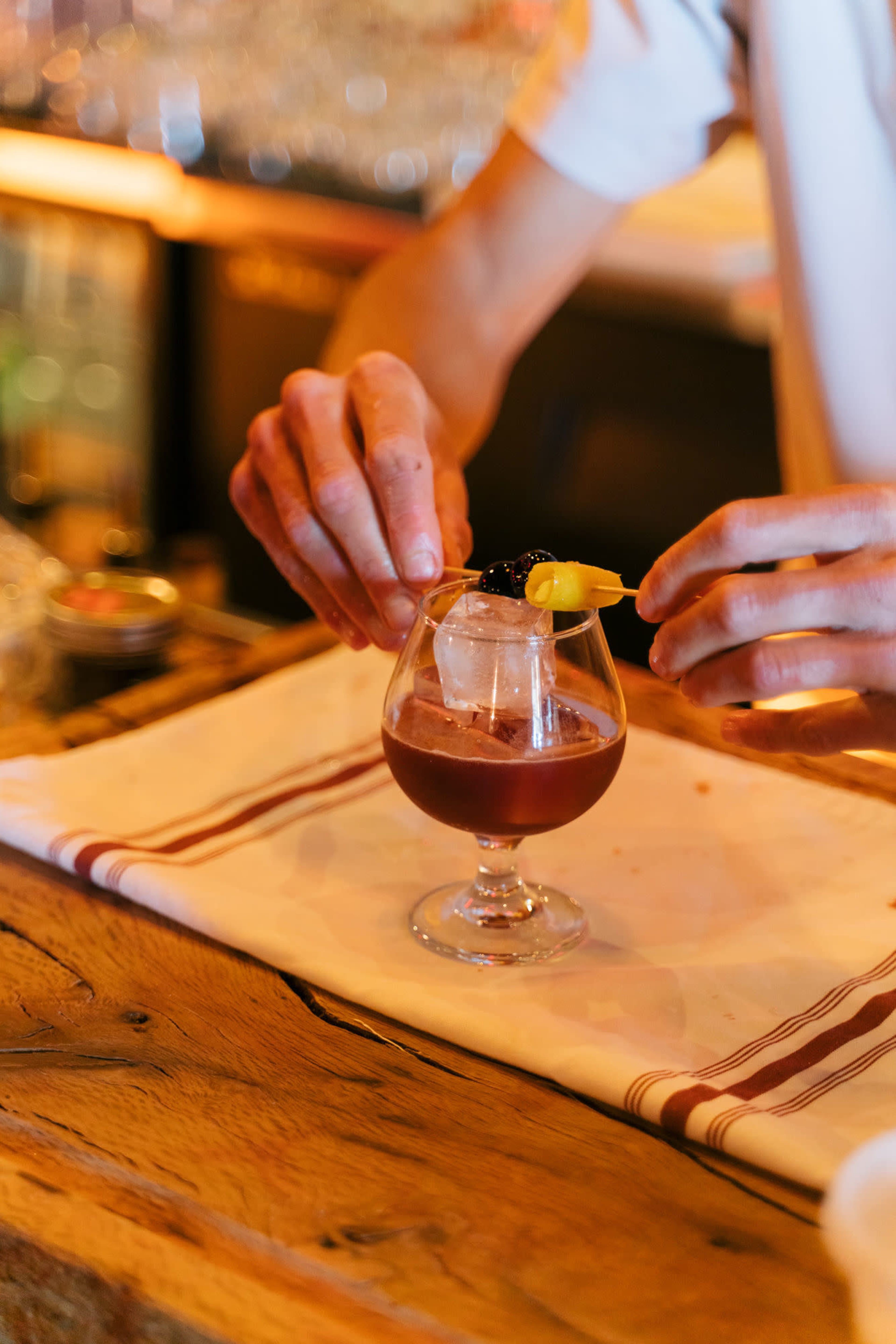 A bartender adds a garnish of a twist of citrus and a cherry to a cocktail in a glass.